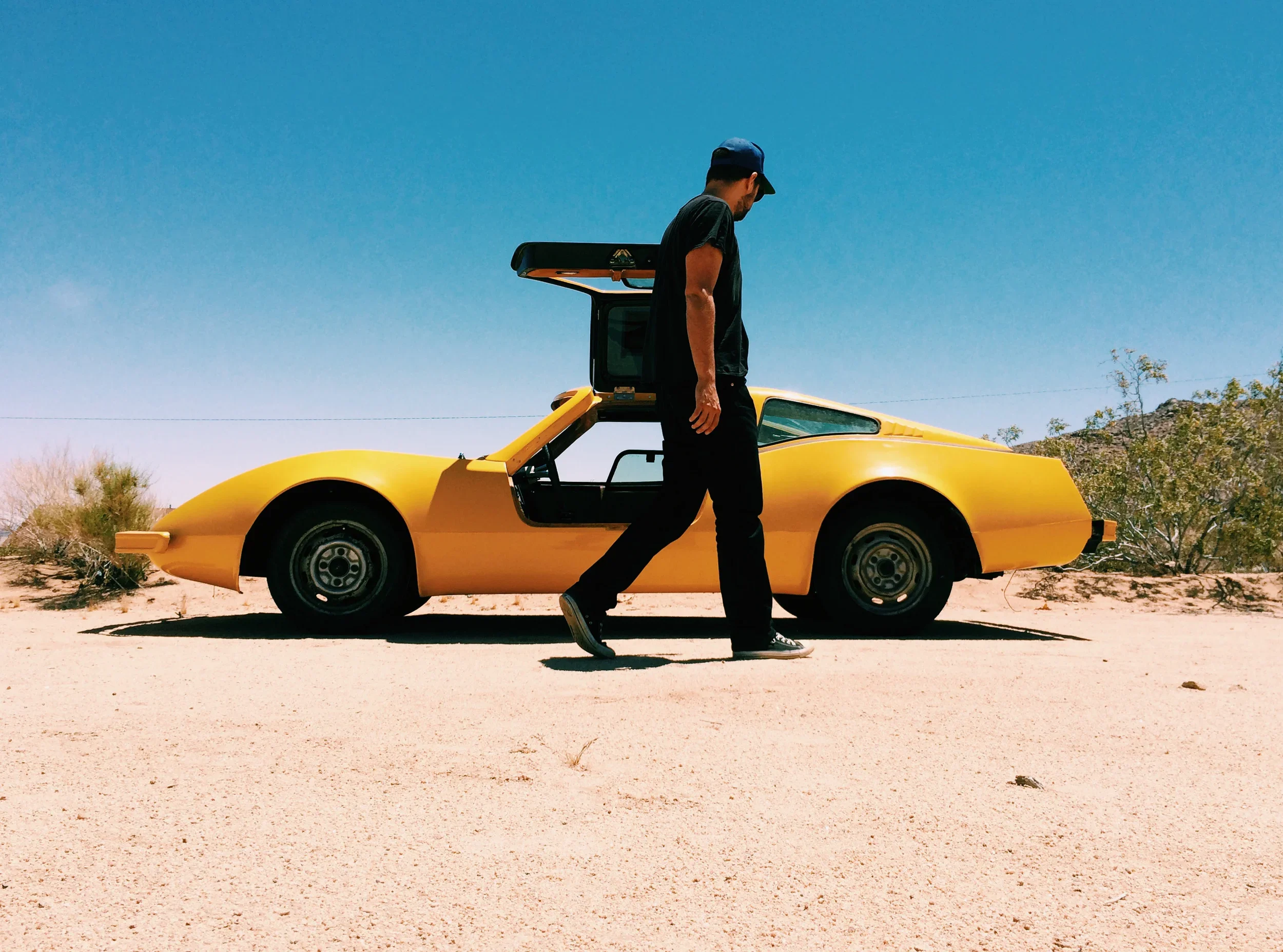 Man walking in front of a yellow sports car with butterfly doors open in a desert landscape under a clear blue sky.
