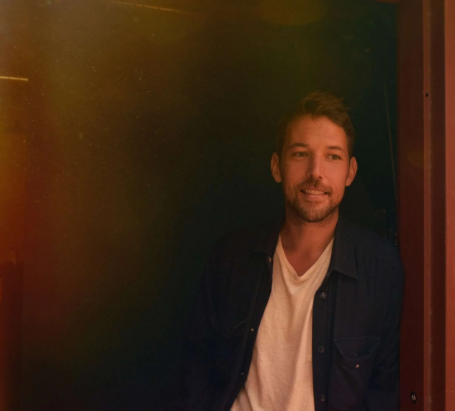 A young man with brown hair and a beard, smiling, wearing a black jacket over a white shirt, standing in a dark corner.