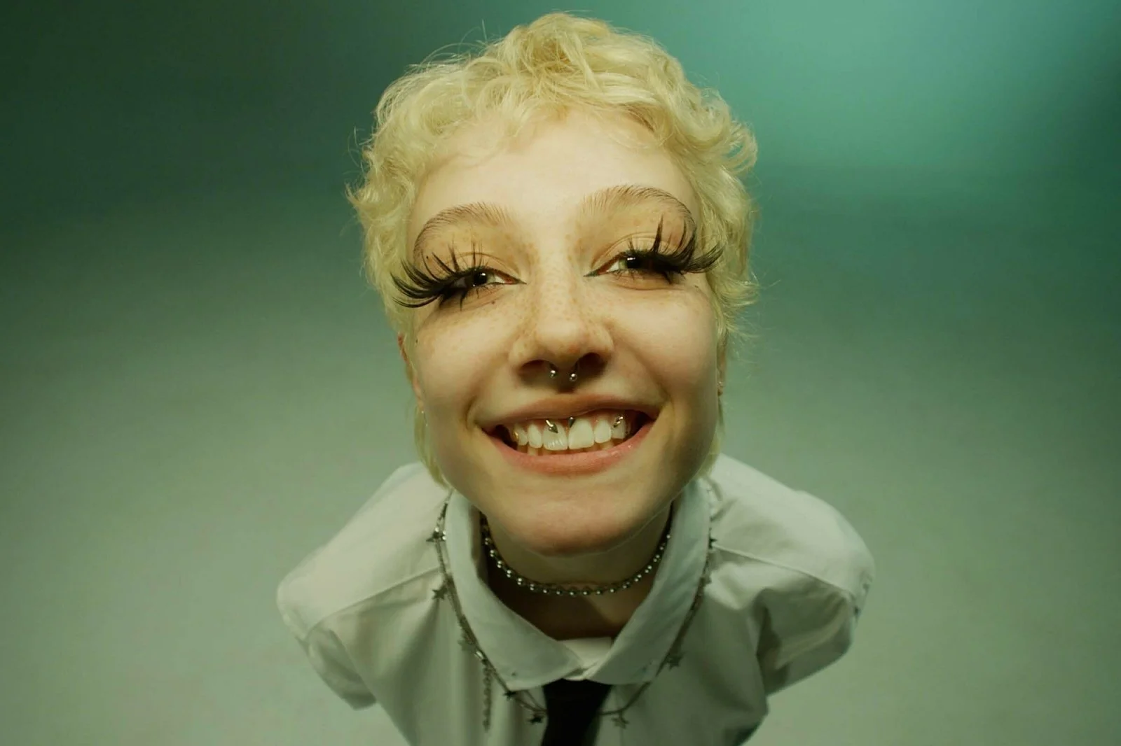 Close-up of a smiling person with short, curly blonde hair, wearing large false eyelashes, a septum piercing, a choker necklace, and a studded shirt against a plain green background.