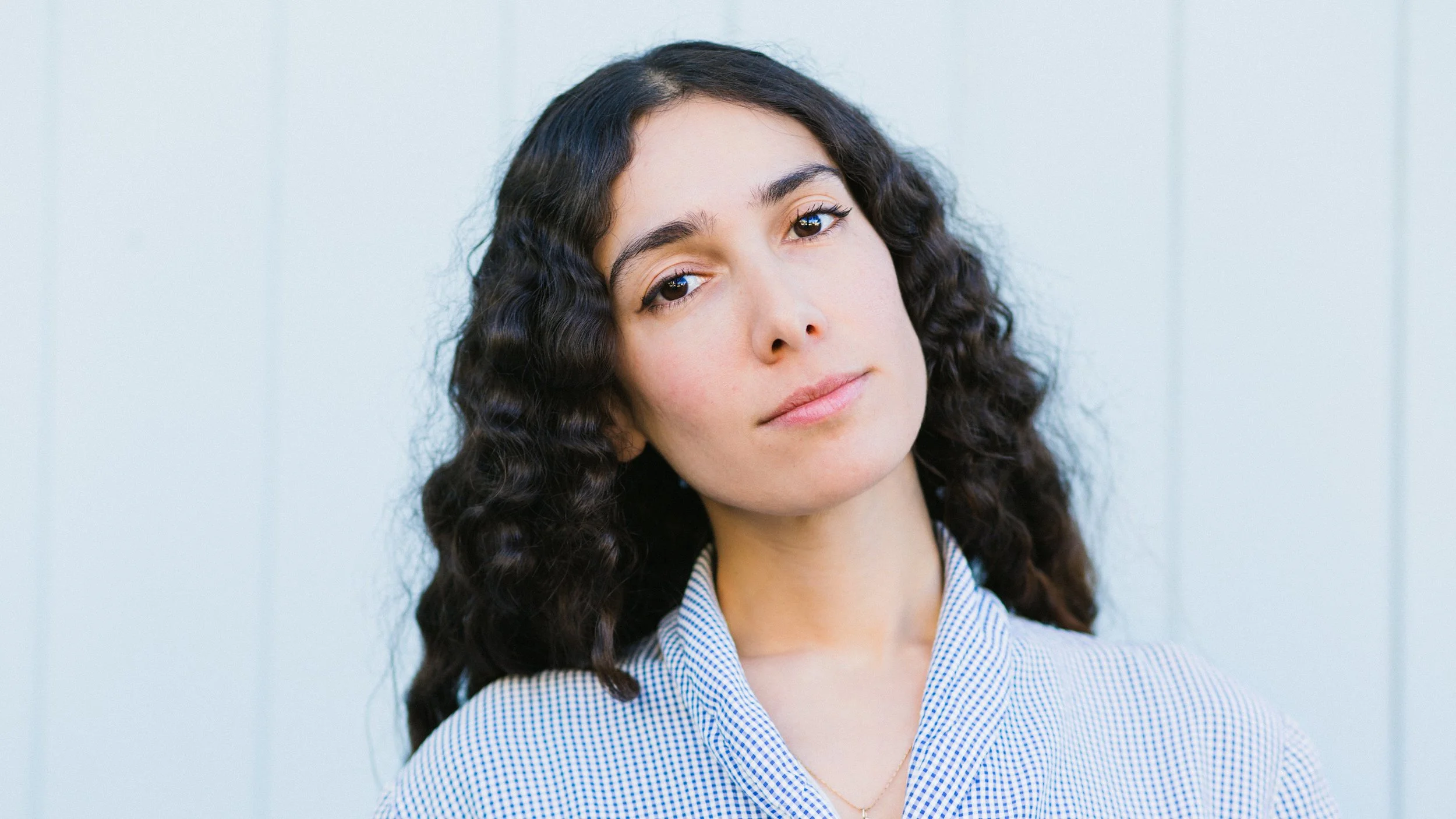 A woman with long, dark, curly hair wearing a gray and white checkered shirt, standing against a light-colored wall.
