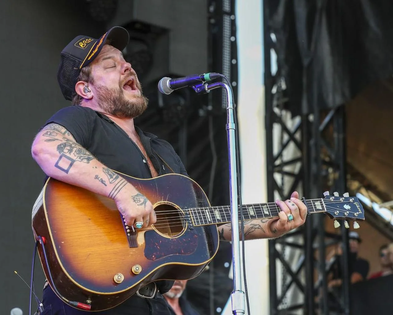 A male musician with tattoos on his arms, wearing a black shirt and a black cap, passionately singing and playing an acoustic guitar on stage during a performance.