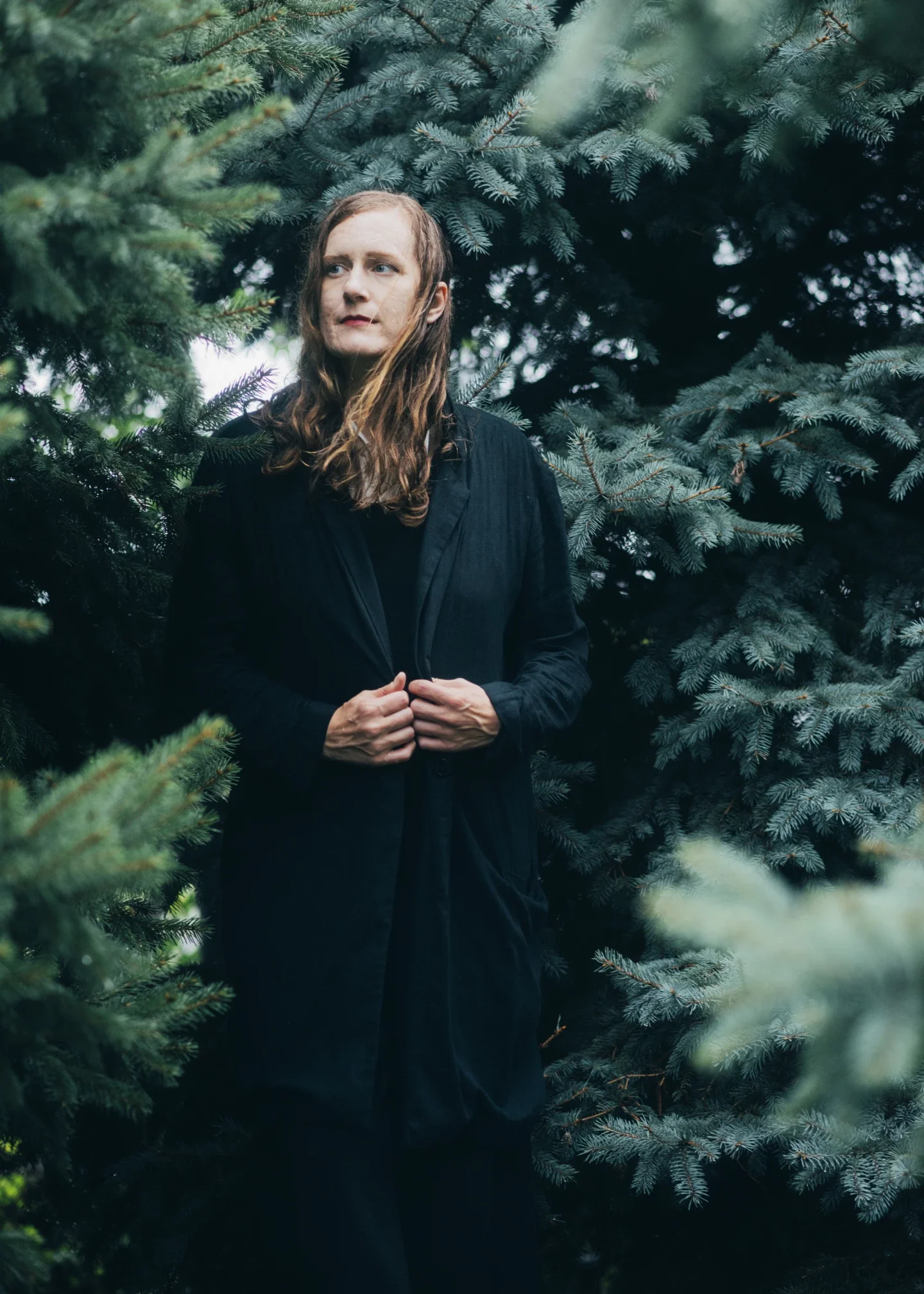 A woman with long, wavy brown hair standing amidst dense green pine trees, wearing a long black coat, with her hands clasped in front of her.