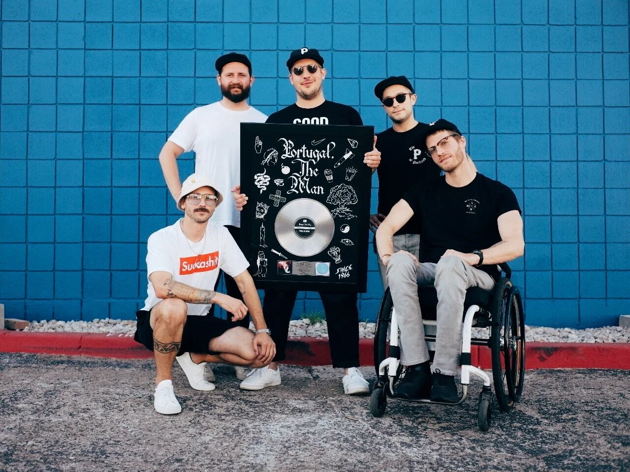 Group of five young men posing outside against a blue brick wall, with one holding a framed record of Portugal. The Man's album, and one in a wheelchair. All wear casual clothing and sunglasses.