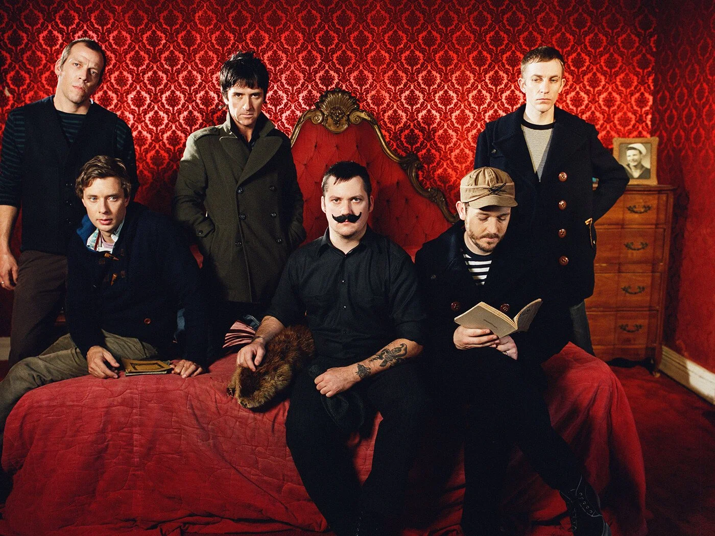 Group of seven men in vintage-style clothing posing in a room with red patterned wallpaper, some sitting and some standing, one holding a book, one wearing a fake mustache, and another with a tattoo sleeve.