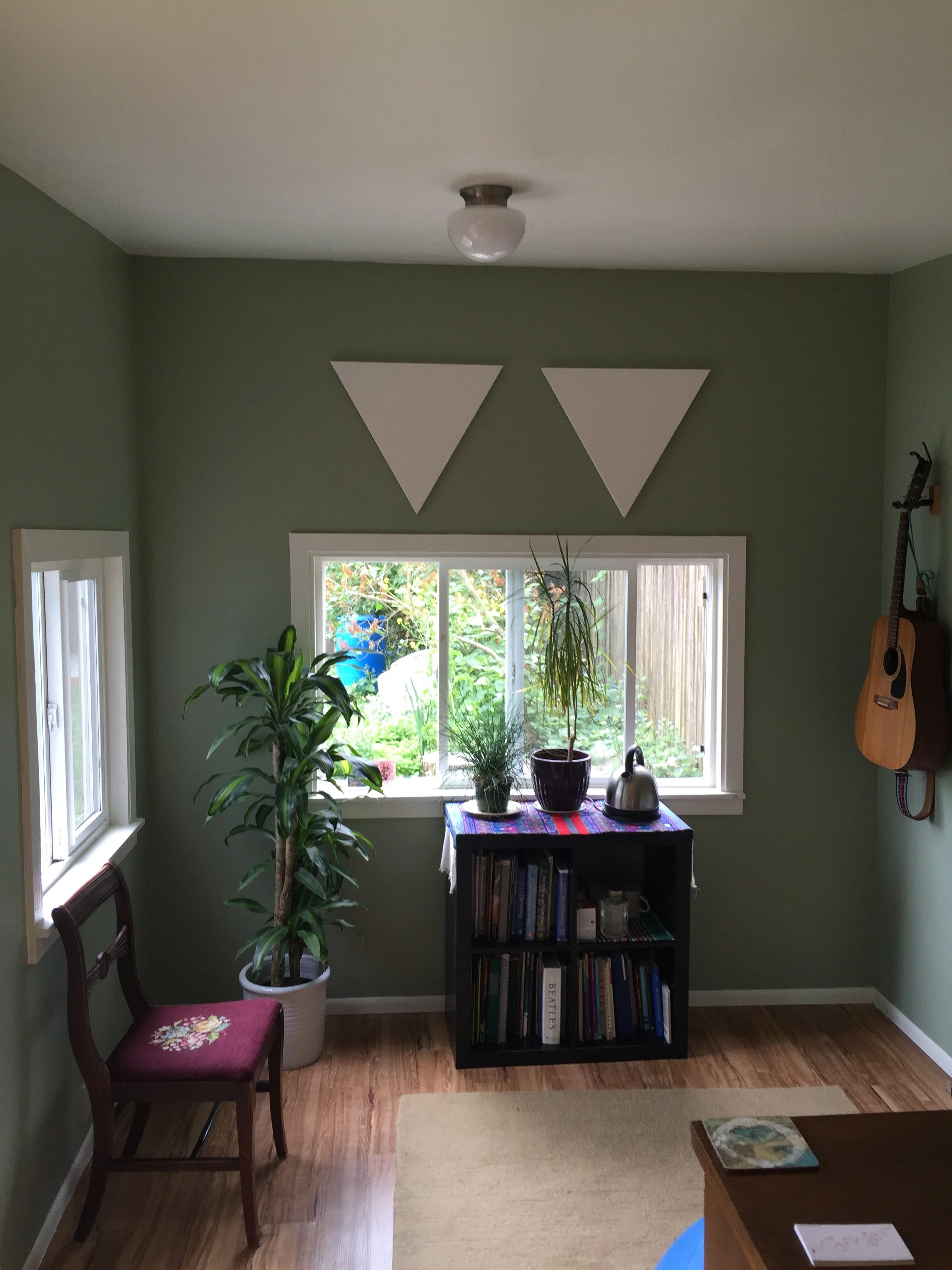 Living room with green walls, a window with potted plants on the sill, a bookshelf, a wooden chair with a floral embroidered cushion, a guitar hanging on the wall, and a small table with books and plants.