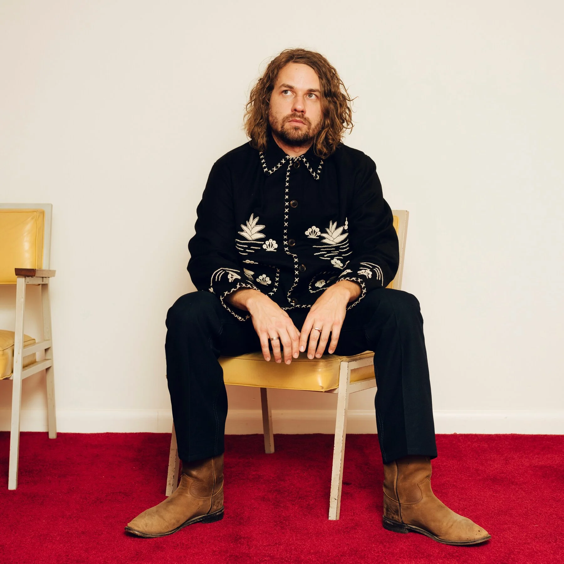 A man with long curly hair and a beard sitting on a yellow chair on a red carpet against a plain white wall, wearing a black embroidered jacket, black pants, and brown boots.