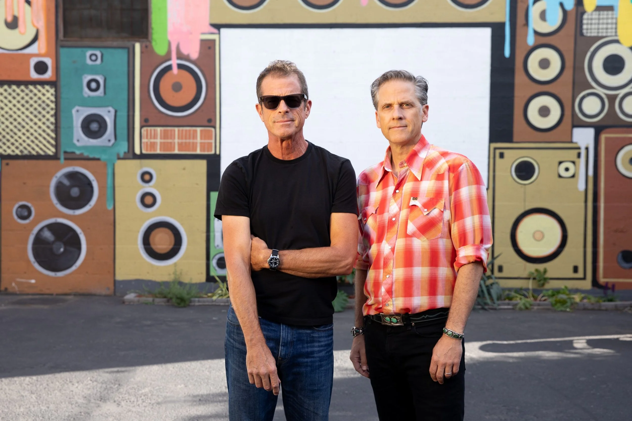 Two men standing in front of a colorful mural of various speakers painted on a wall.