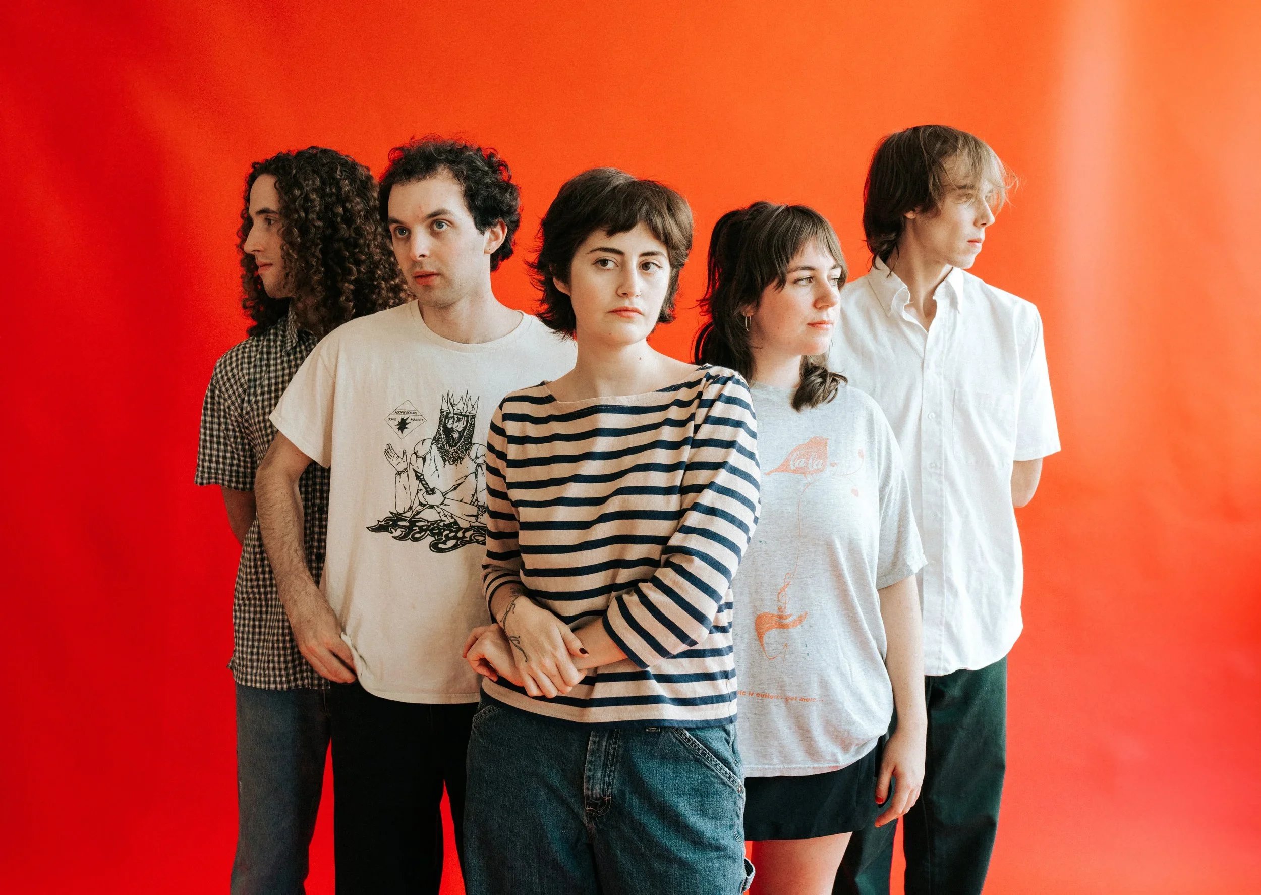 Five people standing in a line against an orange background, looking in different directions, dressed casually.