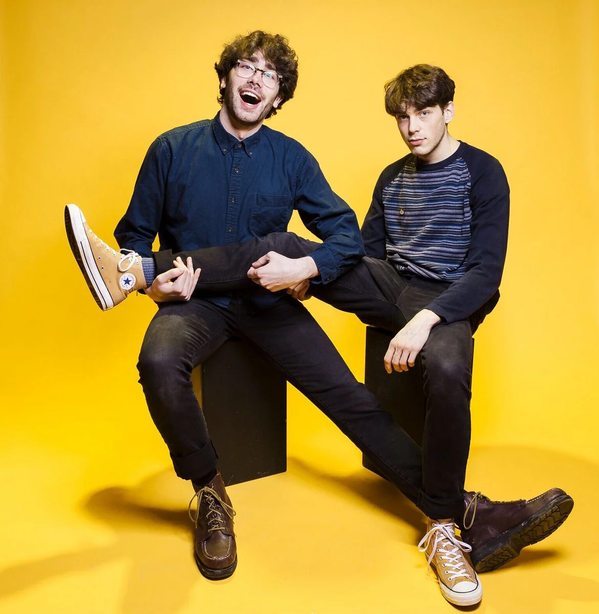 Two young men with dark hair and casual clothing sit against a bright yellow background. One man is holding the other on his lap and smiling. The man being held is looking at the camera with a neutral expression. They are wearing sneakers and jeans.
