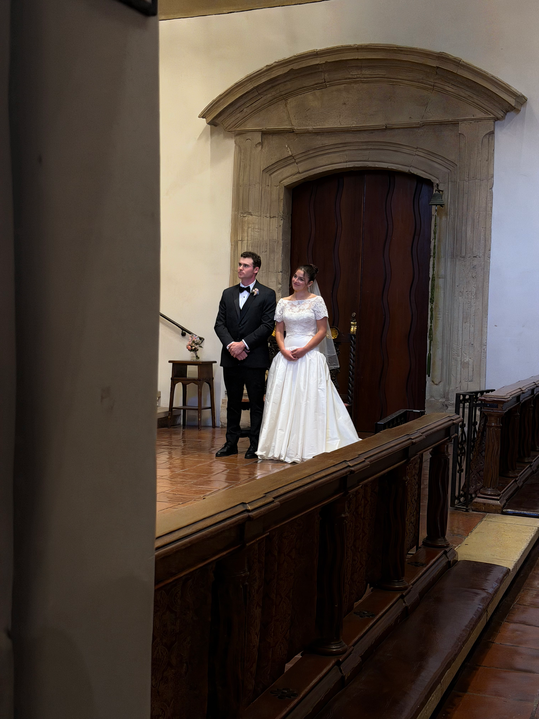 A bride in a white wedding dress and a groom in a black tuxedo standing together at an indoor wedding ceremony.