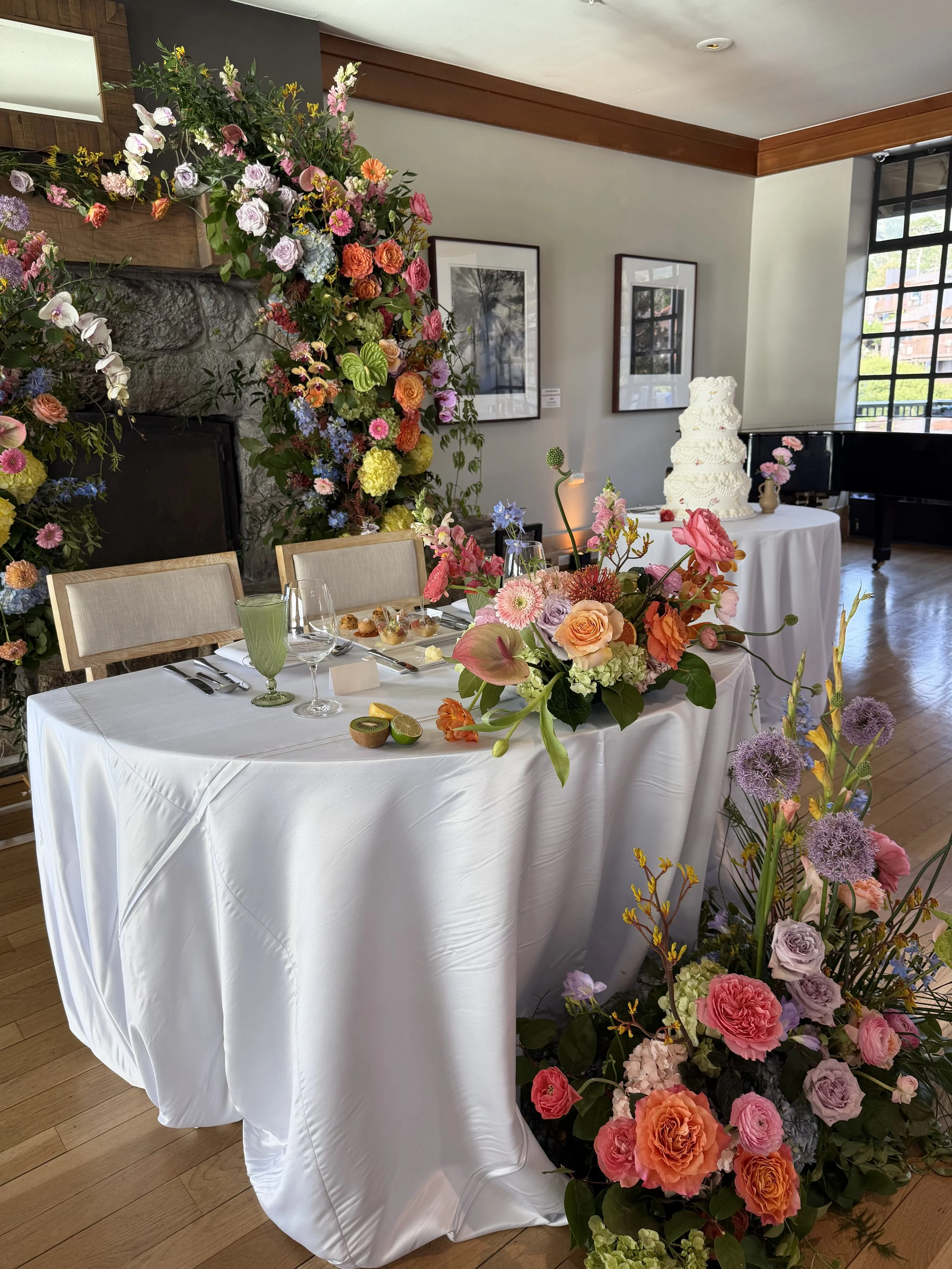 A decorated indoor space for a celebration with a large floral arrangement, a white tablecloth, and a multi-tiered white cake in the background.