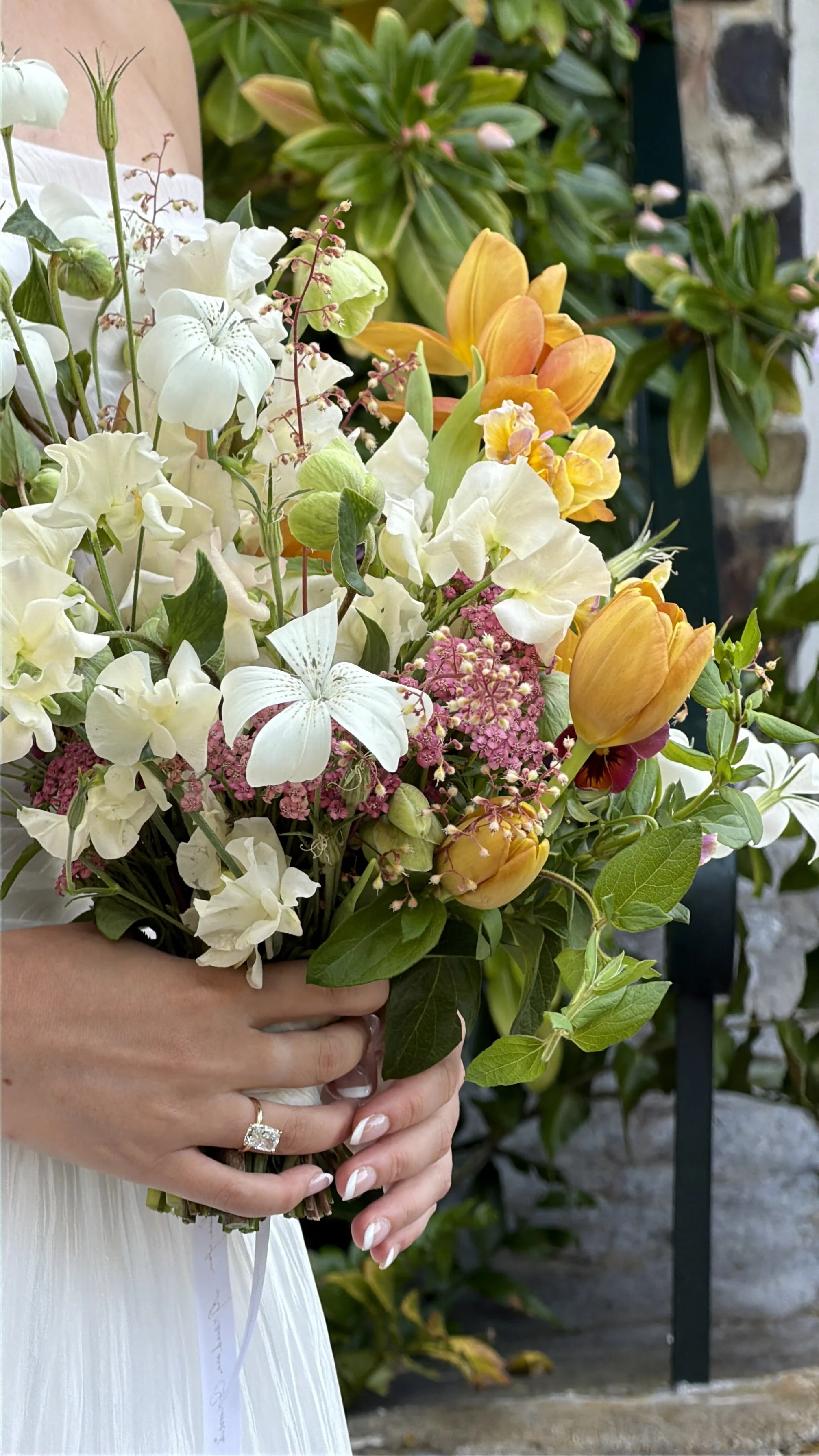 A person holding a bouquet of mixed flowers including white, yellow, and pink blossoms, with a stone and plant background.
