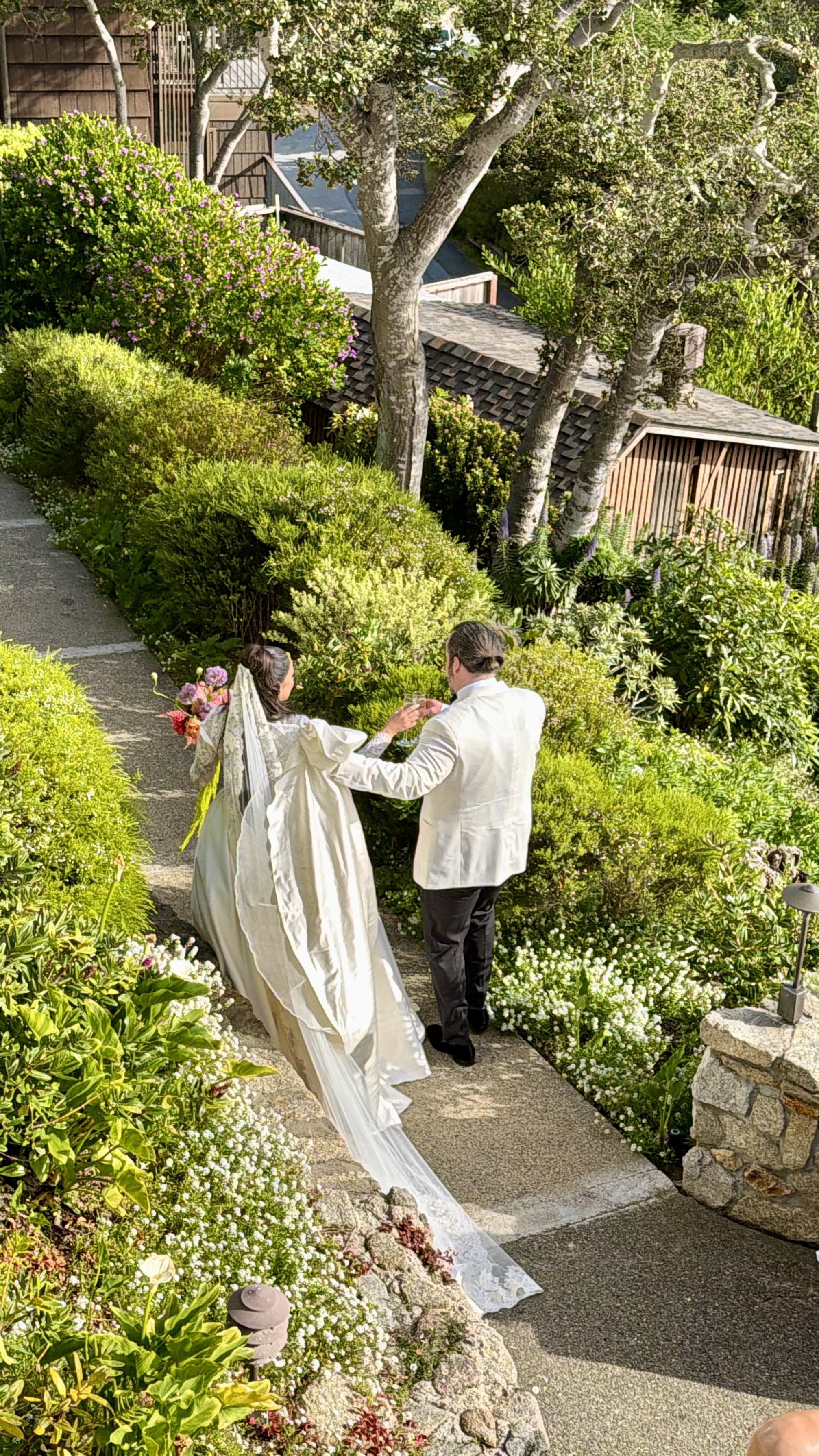 A bride and groom walking together on a garden path, holding hands, amid lush green bushes and flowering plants, with trees and a wooden shed in the background.
