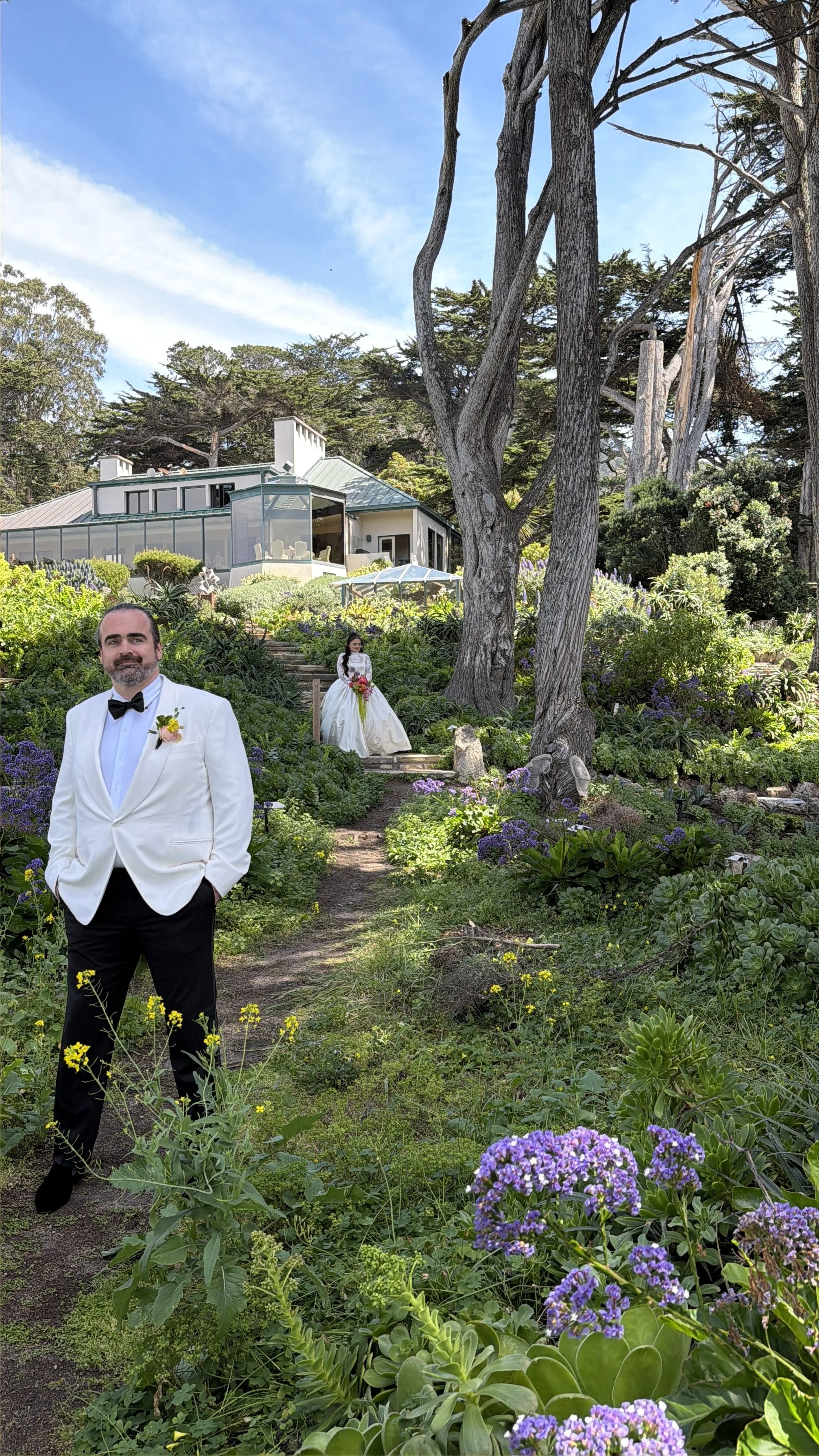 A groom in a white tuxedo with a bow tie and boutonniere, standing outdoors on a garden path, with a bride in a white wedding dress holding a bouquet in the background, surrounded by lush greenery and purple flowers, with a modern house and trees in 