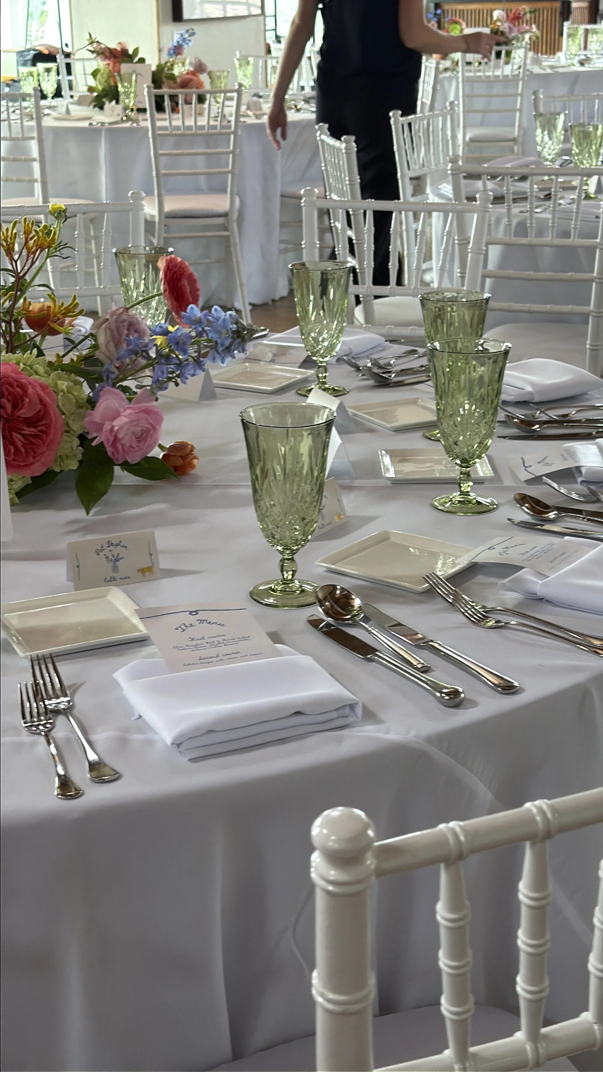 Elegant event table setup with white tablecloths, green glassware, silver cutlery, and floral centerpieces featuring pink and purple flowers. White chairs surround the tables, and a person is seen preparing the decor.