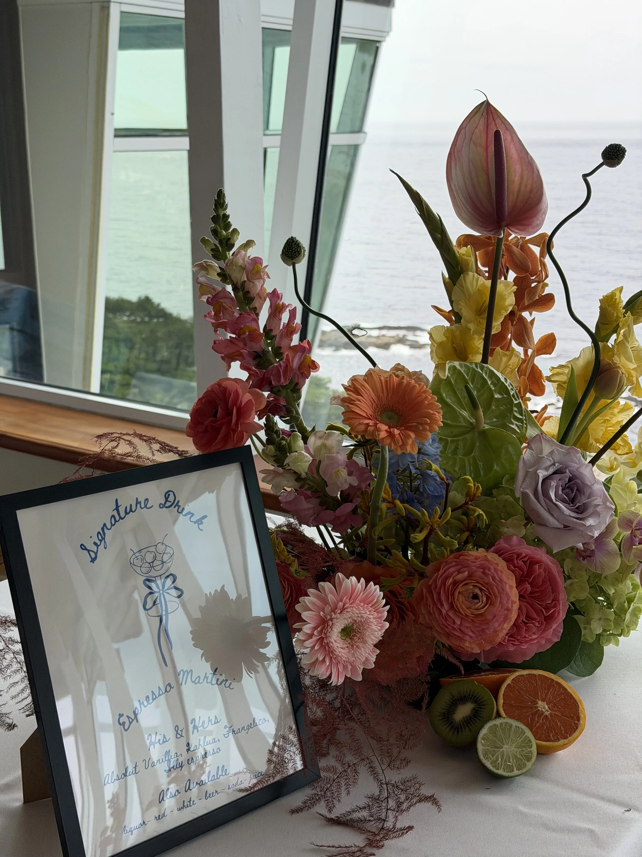 A floral arrangement of pink, orange, yellow, and purple flowers with slices of citrus fruits (orange, lemon, lime, and kiwi) on a table near a window, with a partly visible framed sign in the foreground.