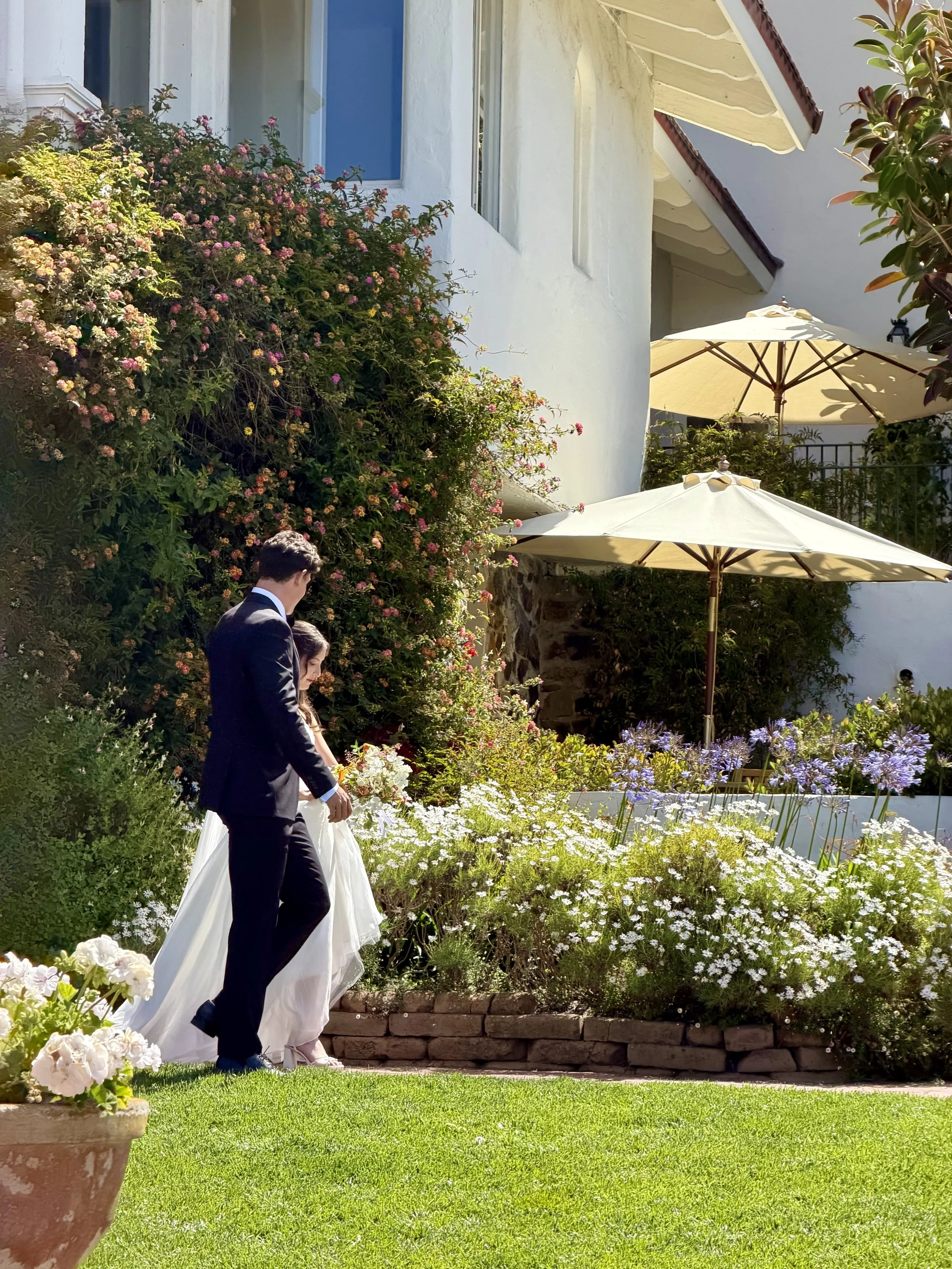 A man and woman dressed in formal wedding attire walking through a garden with blooming flowers, white umbrellas, and a white building in the background.