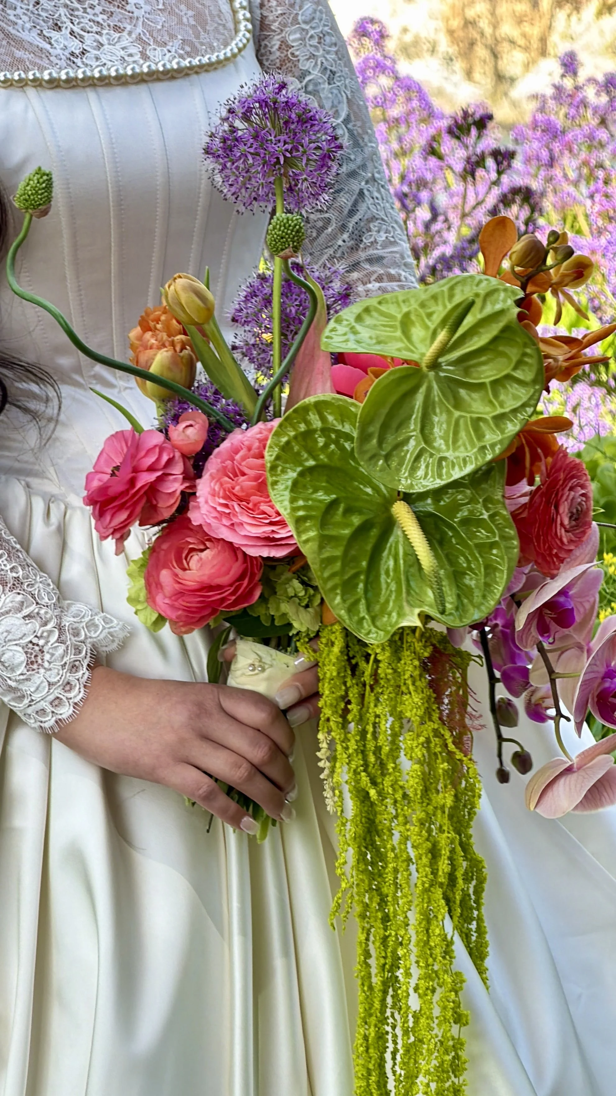 Close-up of a bride in a white dress holding a colorful bouquet of pink roses, purple allium, green Anthurium, orange orchids, and trailing yellow solidago, with her lace sleeve partially visible.