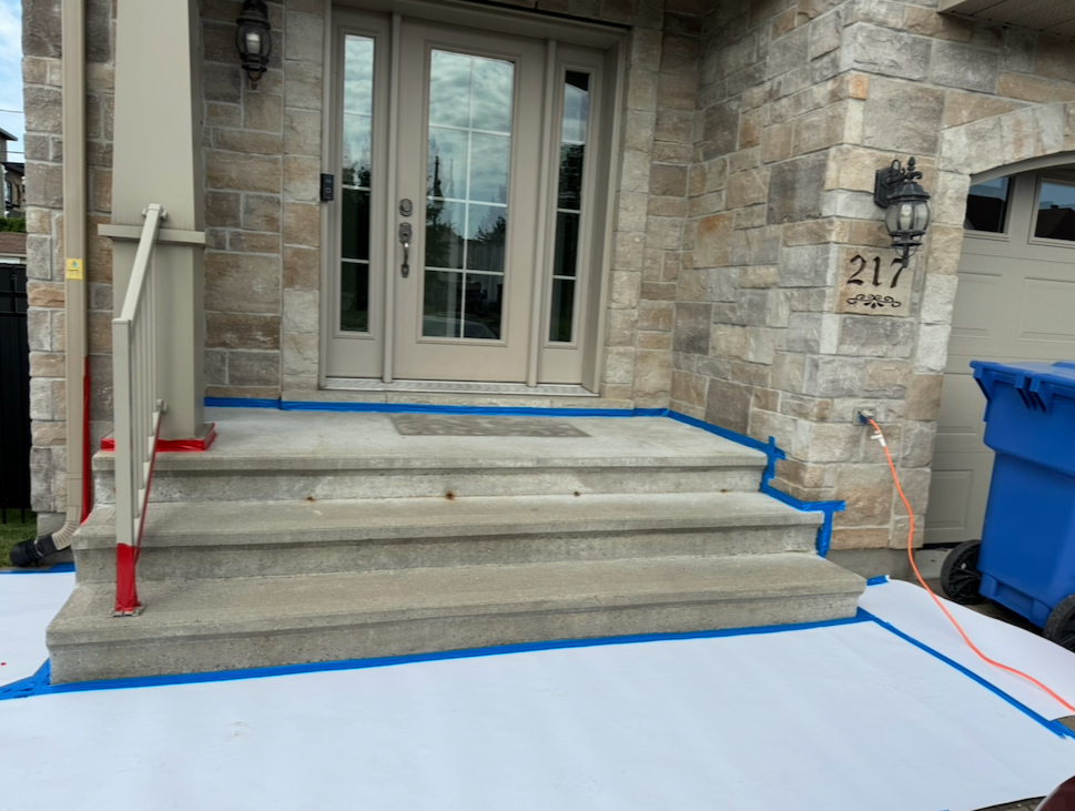 Escalier en béton menant à la porte d'entrée d'une maison en pierre, avec des protections de travaux bleues et un bac de recyclage bleu à côté.