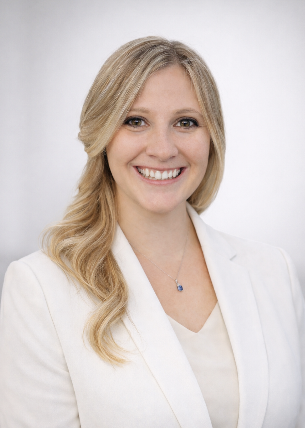 Lindy, a professional with long, curled light-blonde hair, light skin, and a friendly expression. She is wearing a white suit jacket, a white blouse, and a simple necklace against a neutral background.