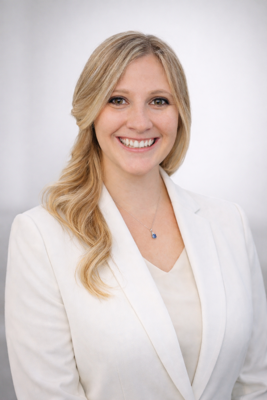 Portrait of Lindy with long blonde hair, hazel eyes, and smiling. Lindy is wearing a white blazer and white blouse with a simple sapphire pendant necklace