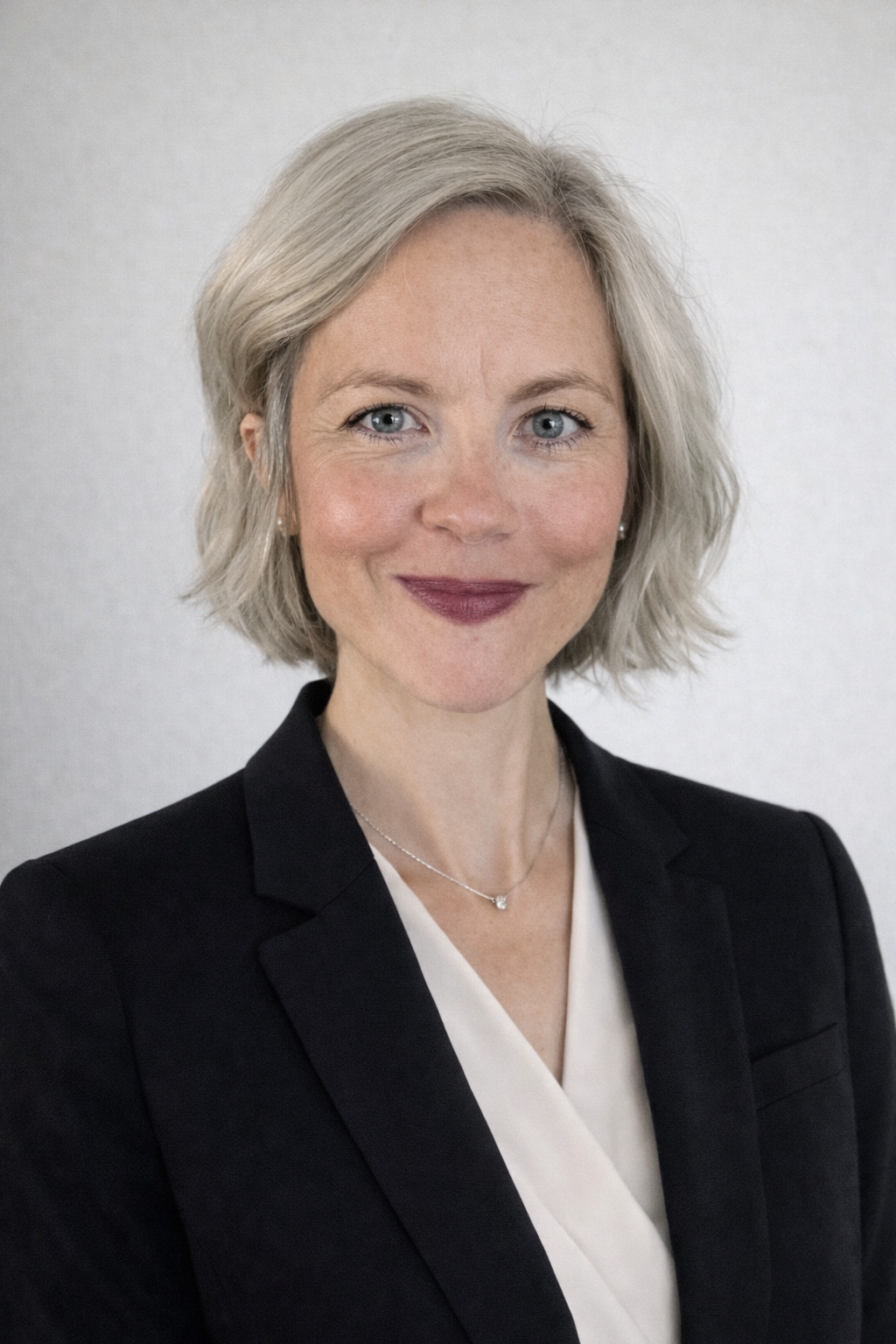 Portrait of Sarah with short light blonde hair, blue eyes, and smiling. Sarah is wearing a dark navy blue blazer and white blouse with a simple diamond pendant necklace