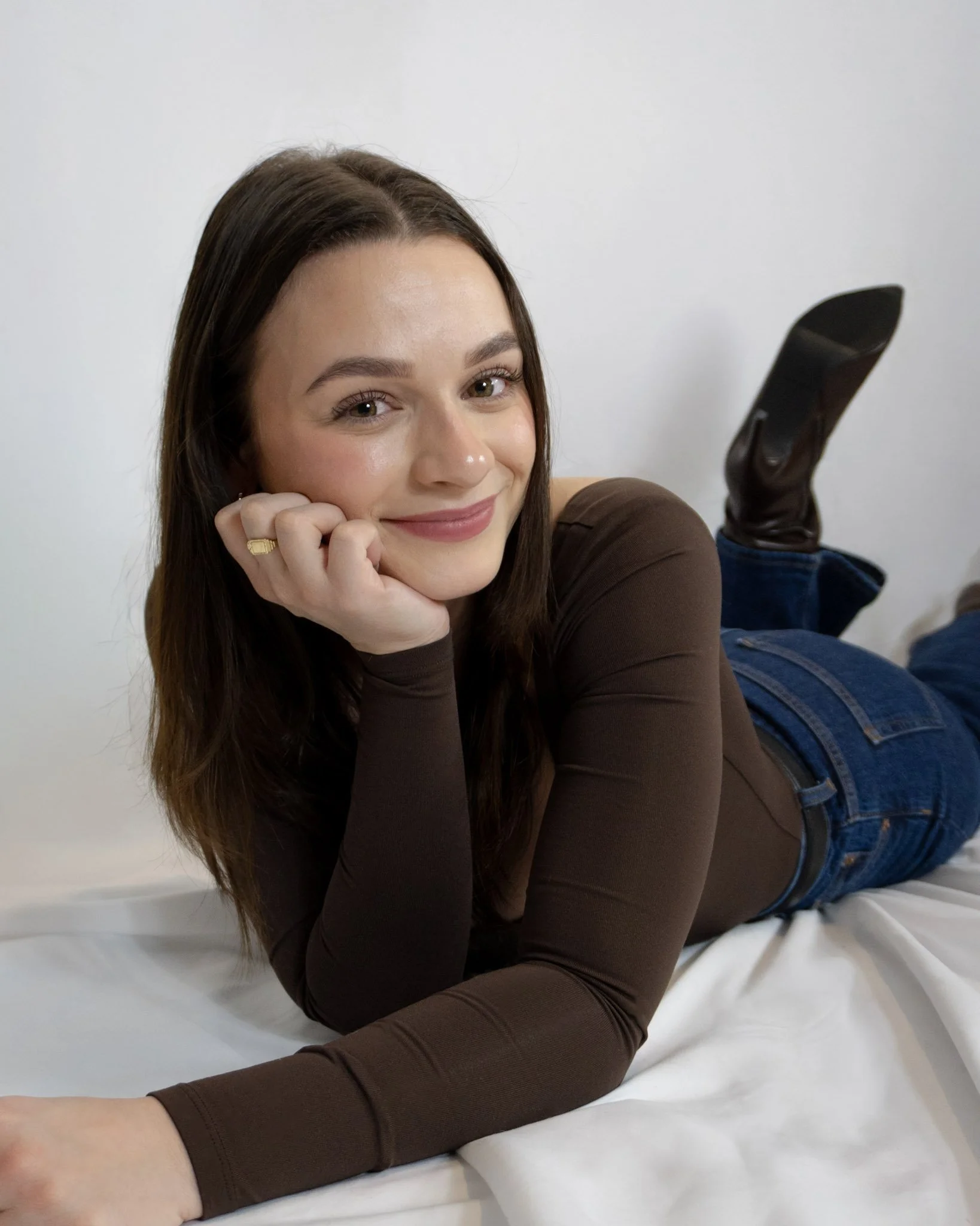 A young woman with long brown hair lying on her stomach on a white bed, smiling and resting her chin on her hand, wearing a long-sleeved brown shirt, dark jeans, and black knee-high boots, with a plain white wall in the background.