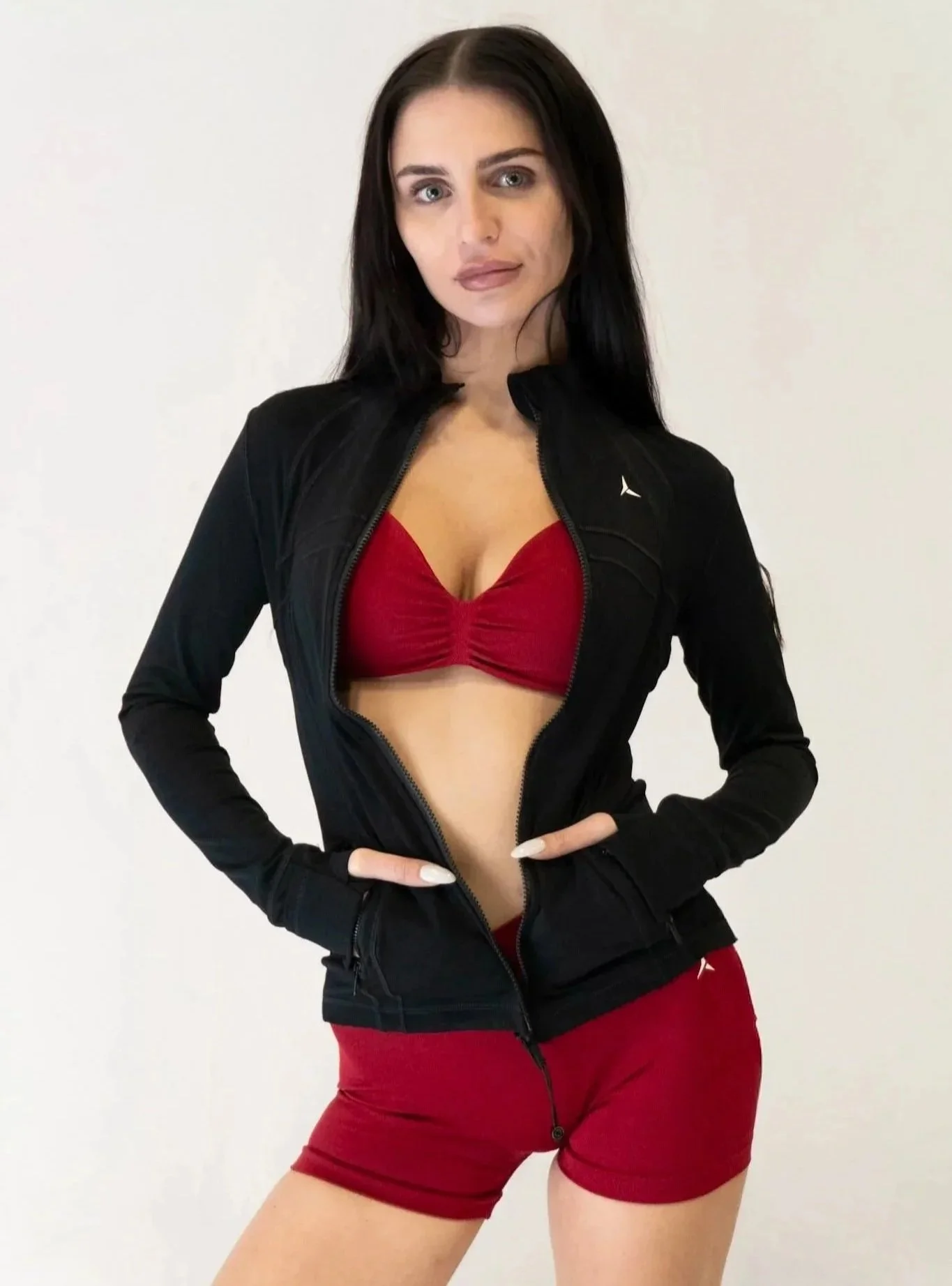 A woman with long dark hair wearing a black jacket over a red sports bra and red shorts, standing against a plain white background.