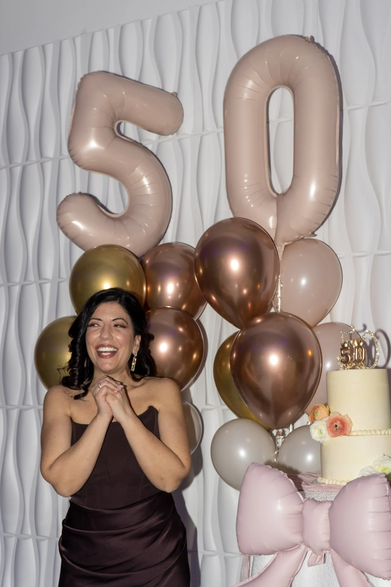 A woman celebrating her 50th birthday, standing in front of a backdrop decorated with gold and pink balloons shaped as the number 50, behind her a white textured wall, with a birthday cake that has a "50" topper and pastel-colored flowers, and a pink