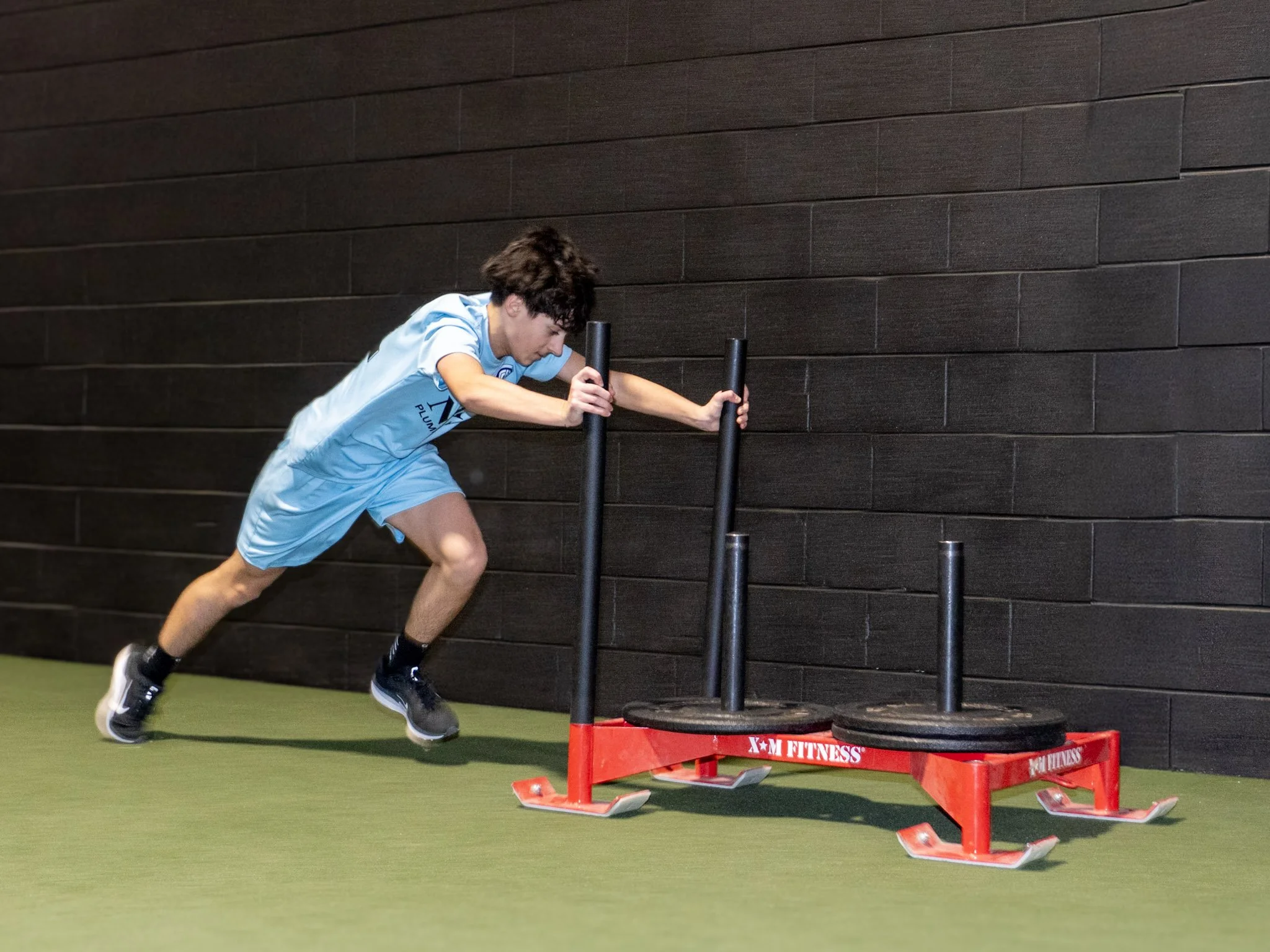 Young man in a light blue workout uniform pushing a sled with weights in an indoor gym with black wall and green floor.
