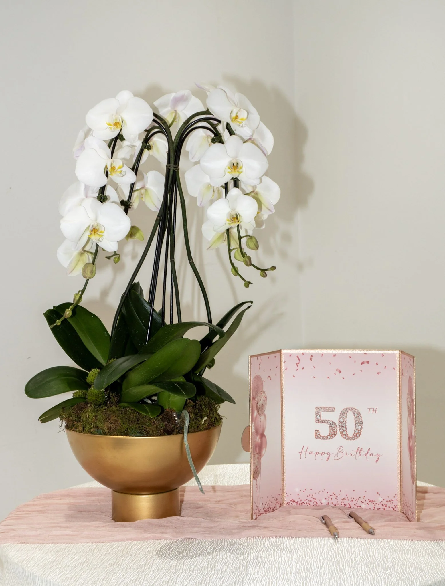 A potted white orchid with multiple blossoms next to a pink birthday card celebrating a 50th birthday, with two pens in front.