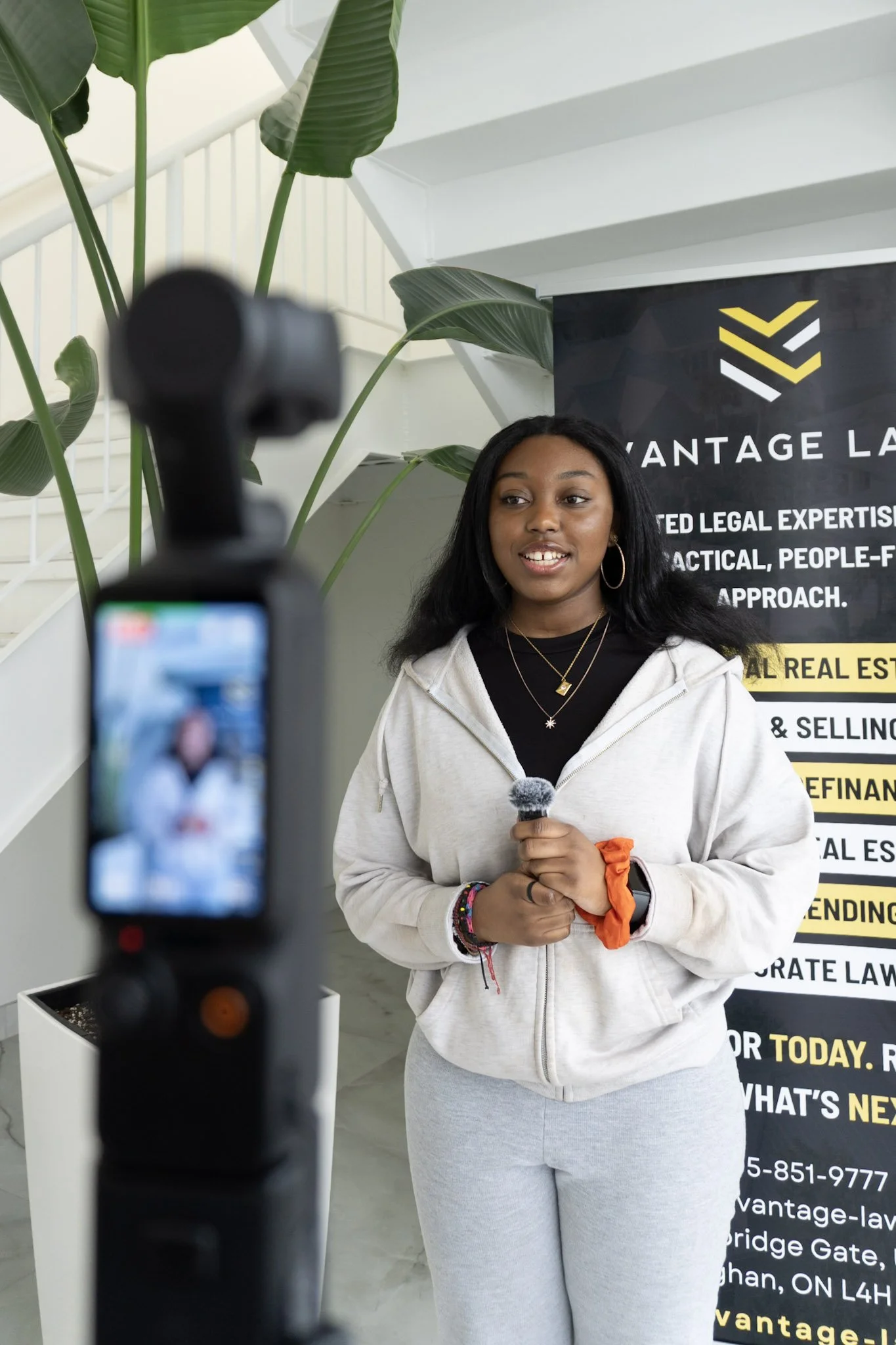 A young woman with long dark hair and hoop earrings speaking into a microphone during a video recording or interview. She's standing in front of a black banner with yellow and white text, and large green plants are visible in the background.