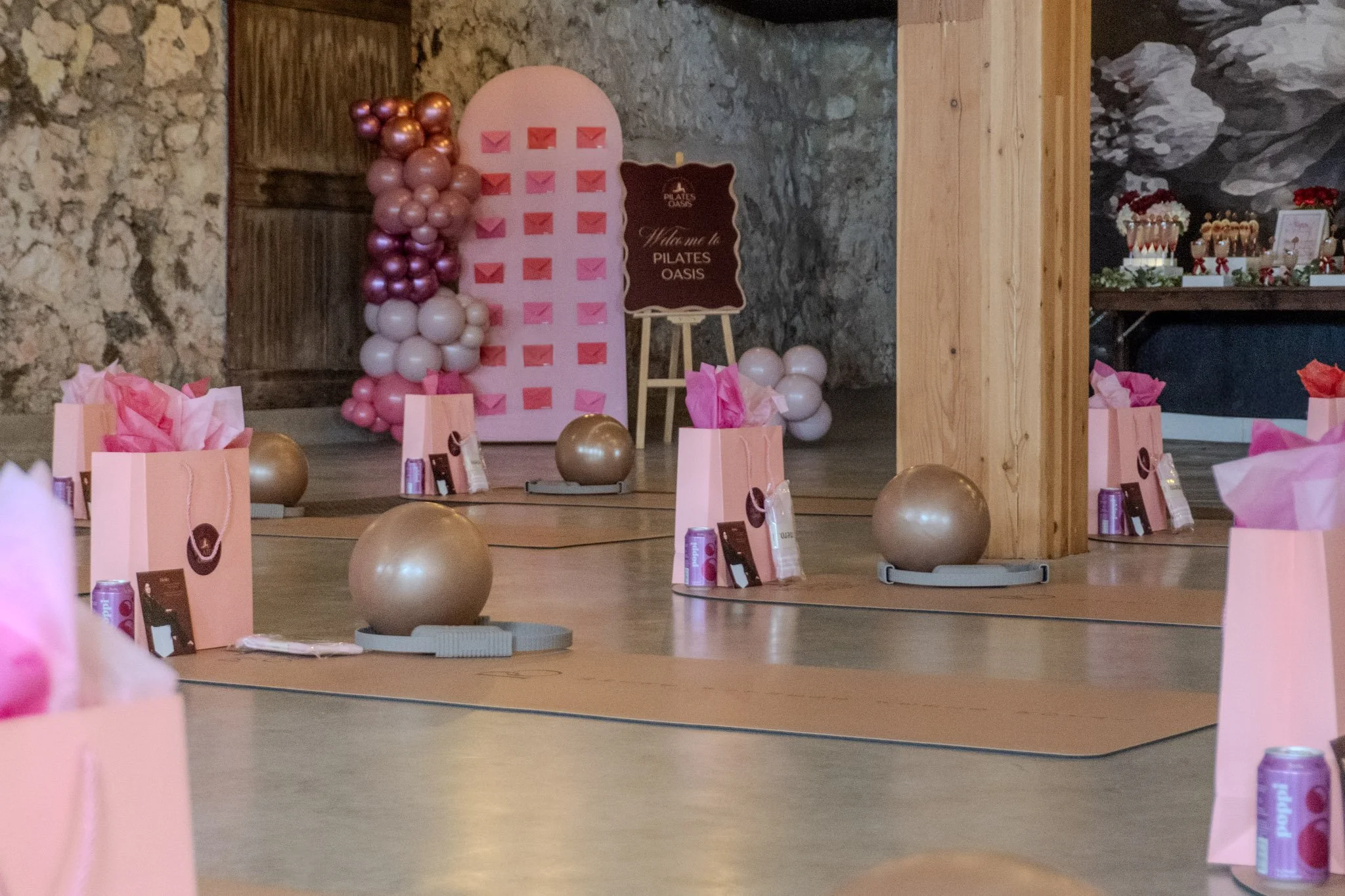 A decorated Pilates studio with pink, gold, and white balloons, pink gift bags with tissue paper, exercise balls, and a festive table, creating a celebratory atmosphere.