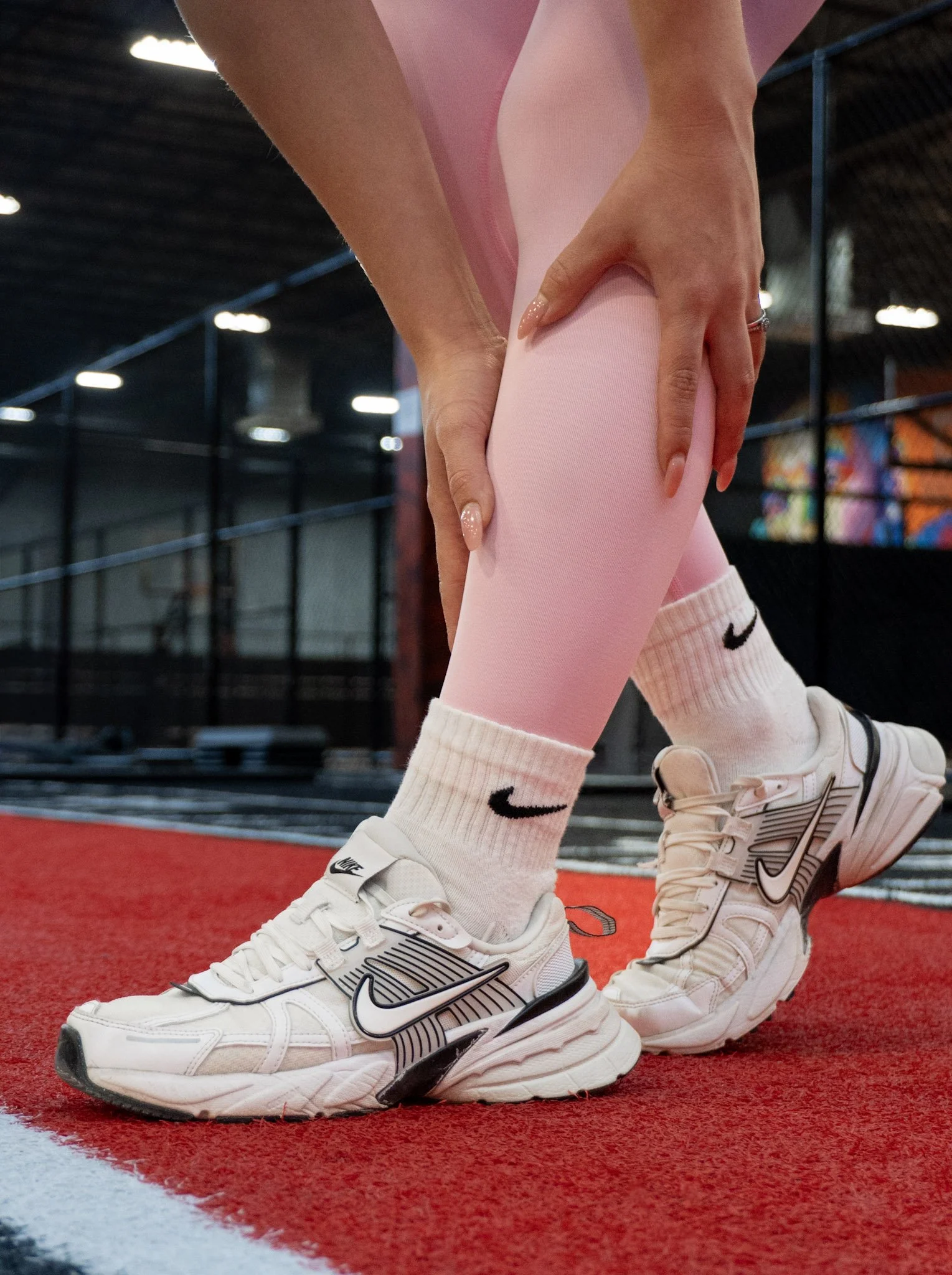 A person wearing white Nike sneakers and pink leggings, bending over and holding their shin on a red indoor track at Mega Gym