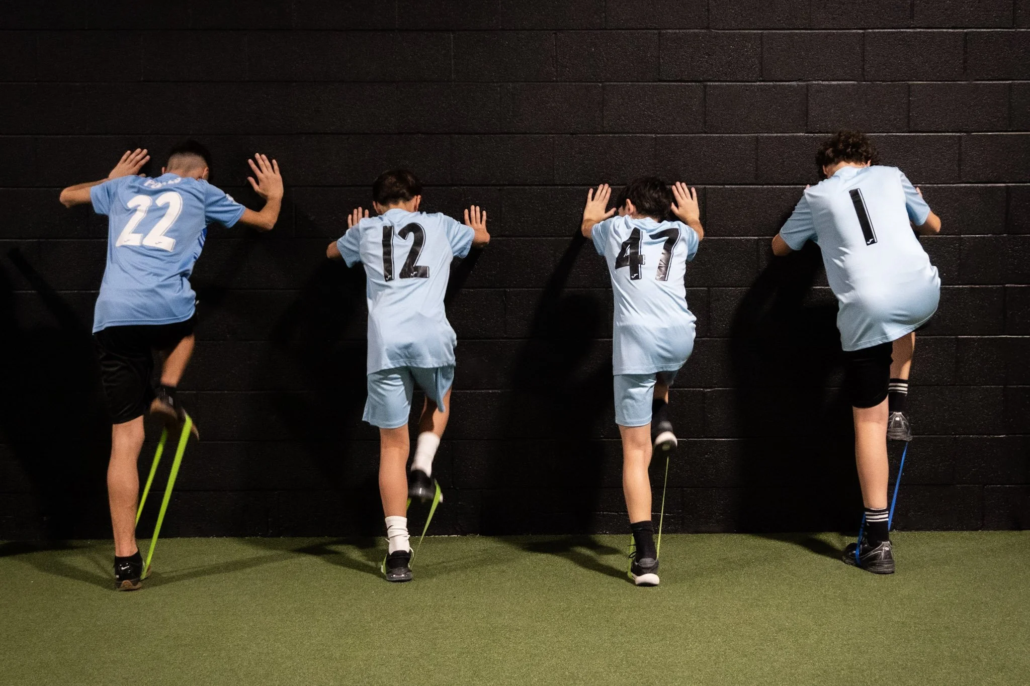 Four young boys in sports jerseys standing against a dark brick wall with one leg raised, facing the wall, with their hands pressed against it.