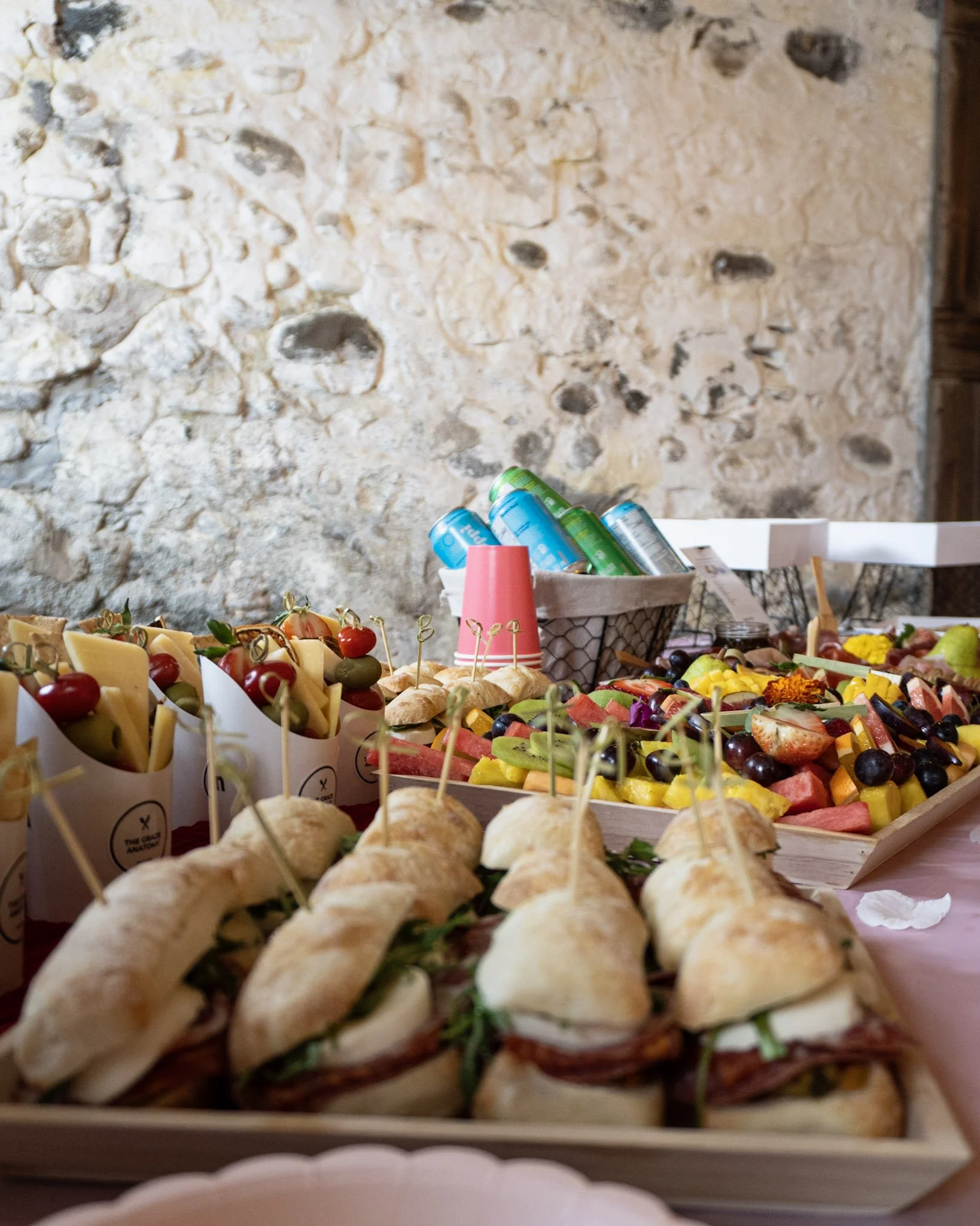 Assorted finger foods including sandwiches, cheese, cherry tomatoes, olives, fresh fruit, and beverages on a table.