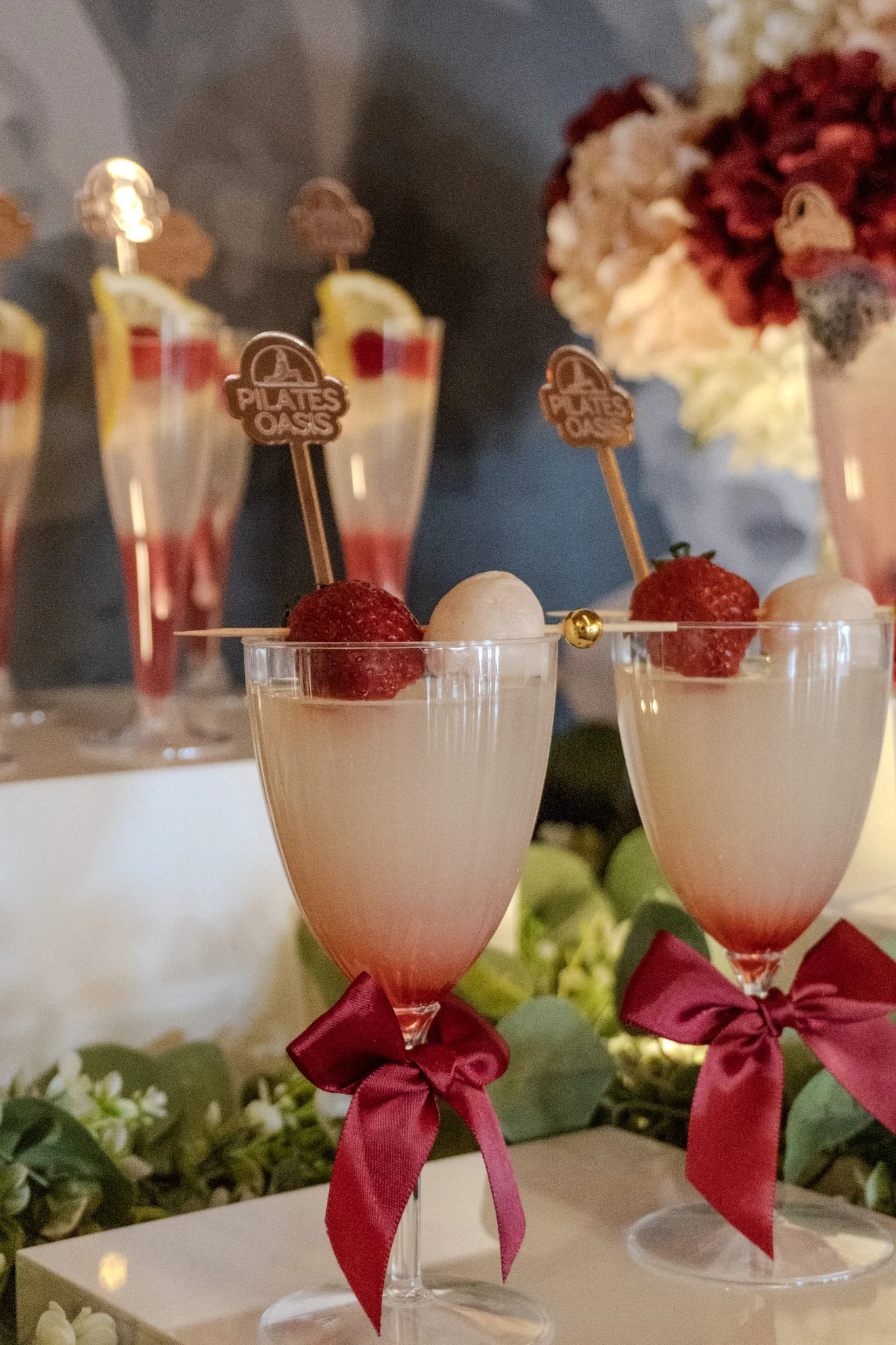 Two champagne glasses filled with a pink cocktail, garnished with strawberries and a white ball, each with a red ribbon tied at the base, on a floral display. In the background, tall glasses with layered drinks, slices of lemon, and pink toppings.