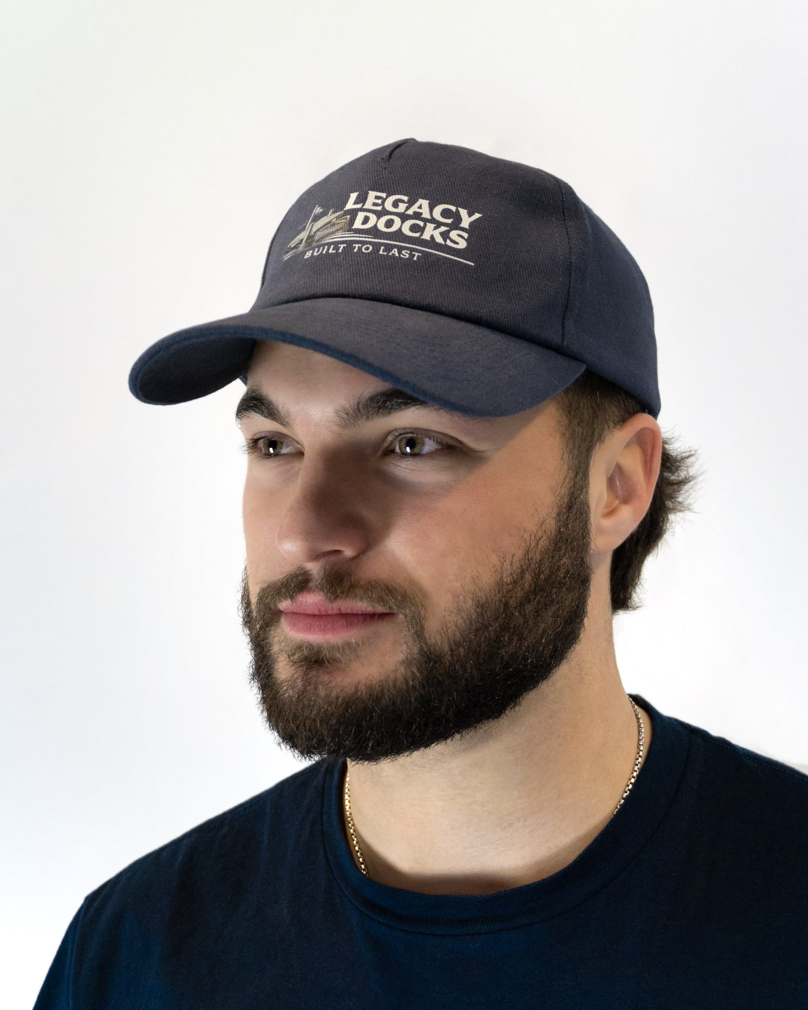 A man wearing a navy baseball cap with a logo that reads "Legacy Docks" and a black T-shirt, standing against a plain white background.