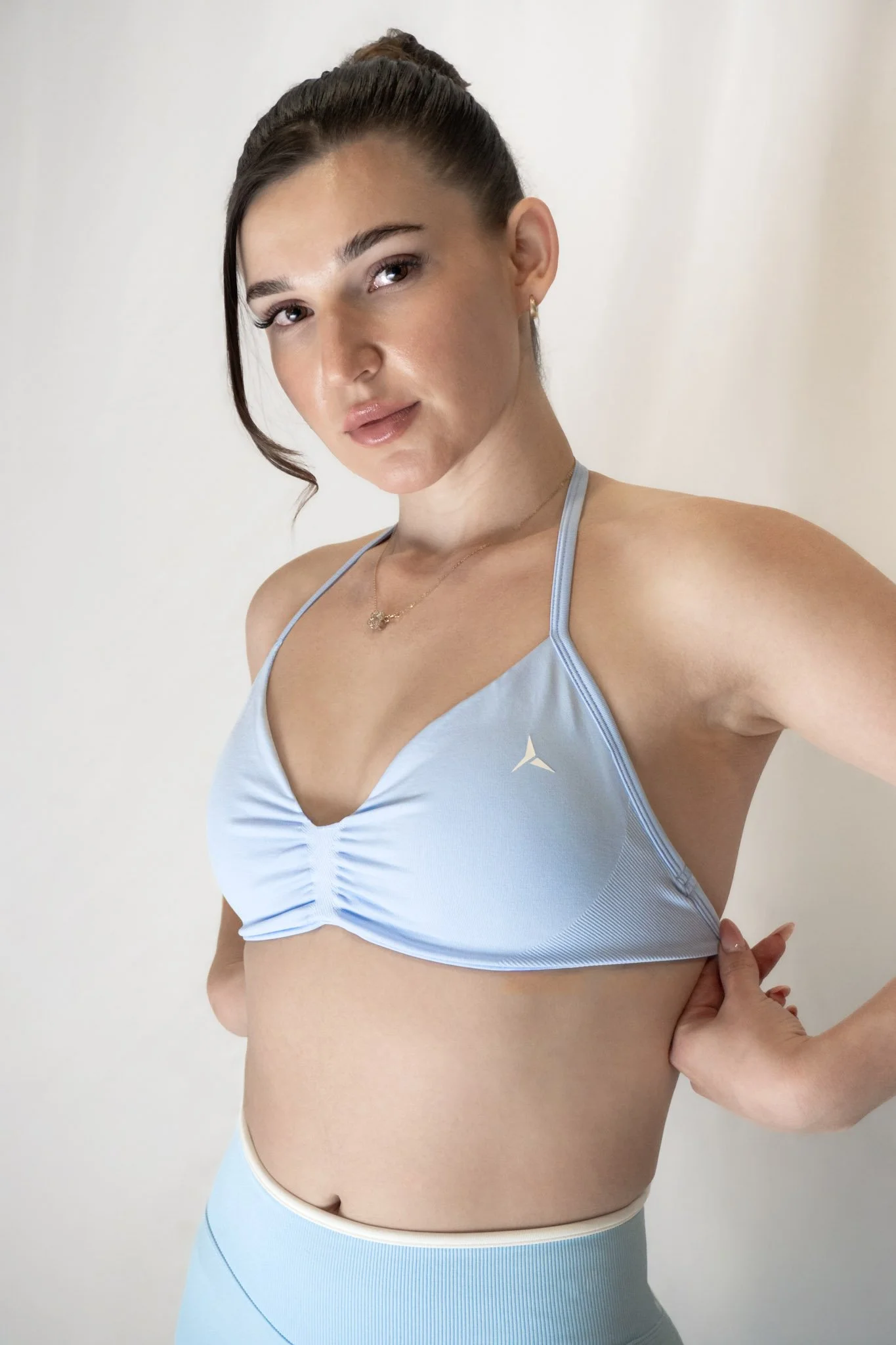 A woman in a light blue athletic sports bra and matching leggings, with her hair in a bun, standing against a plain white wall.