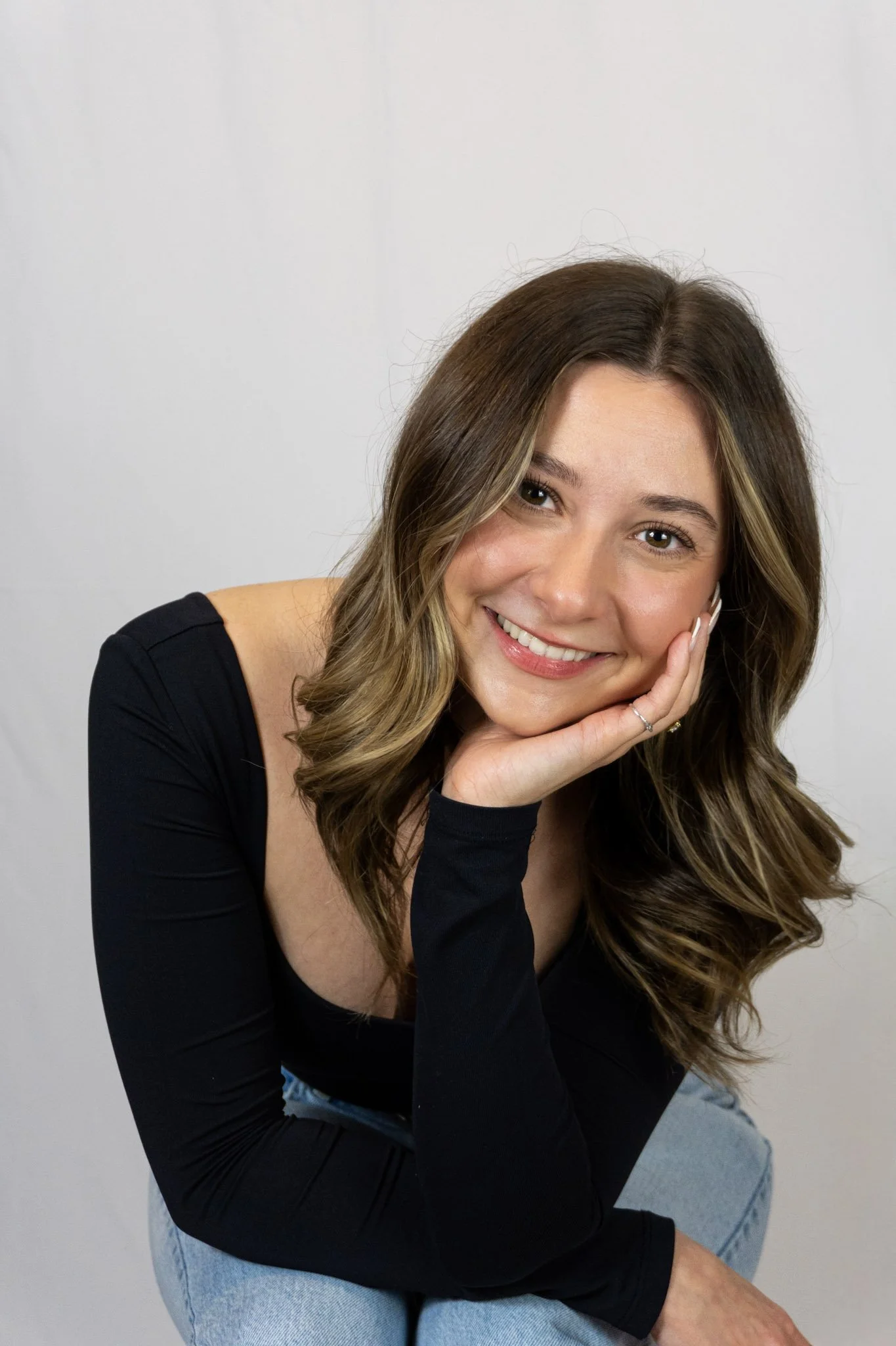 A woman with wavy brown hair and a black top, smiling and resting her face on her hand