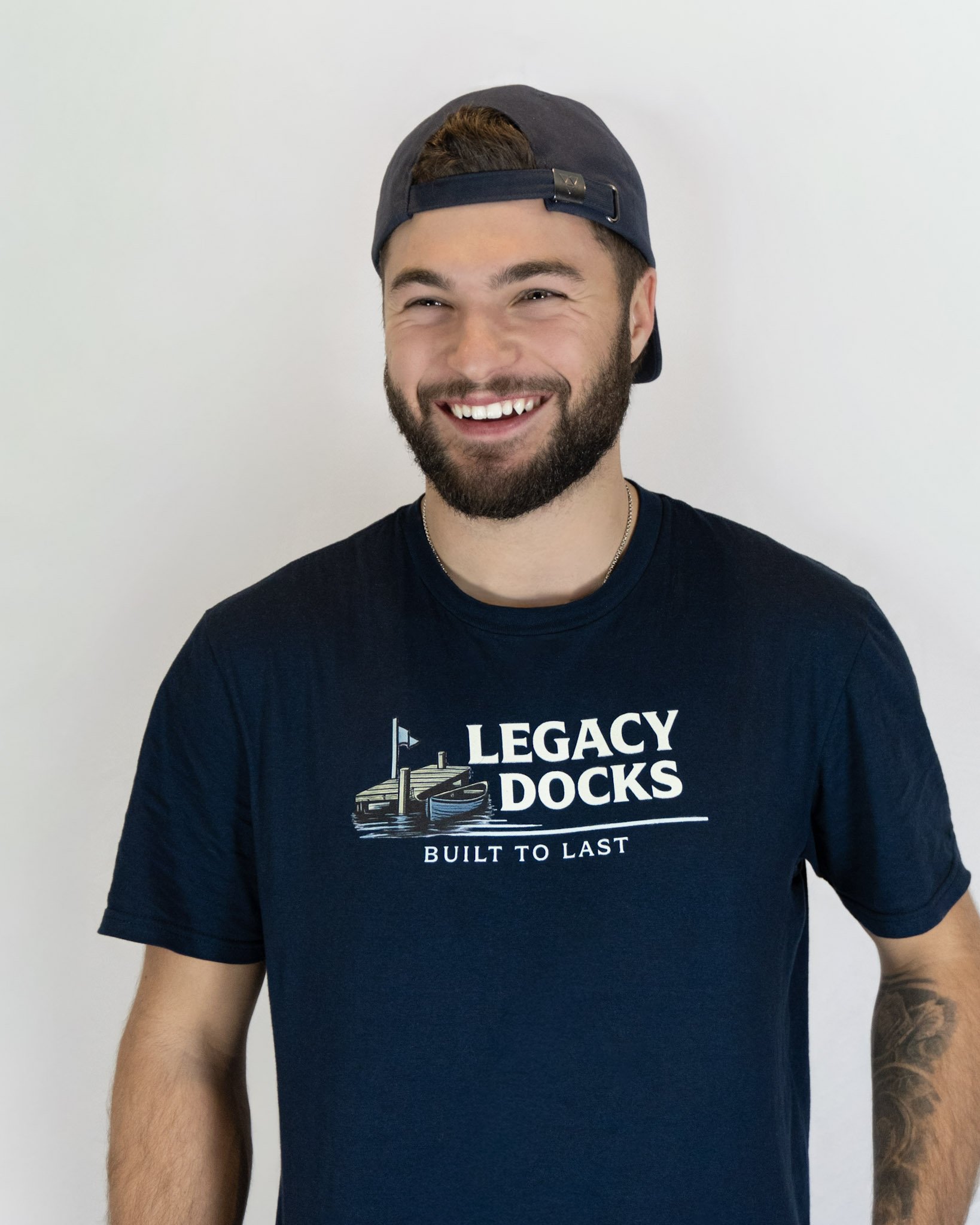A smiling man wearing a dark baseball cap backwards, a black T-shirt with 'Legacy Docks Built to Last' and a dock illustration, stands in front of a plain white background.