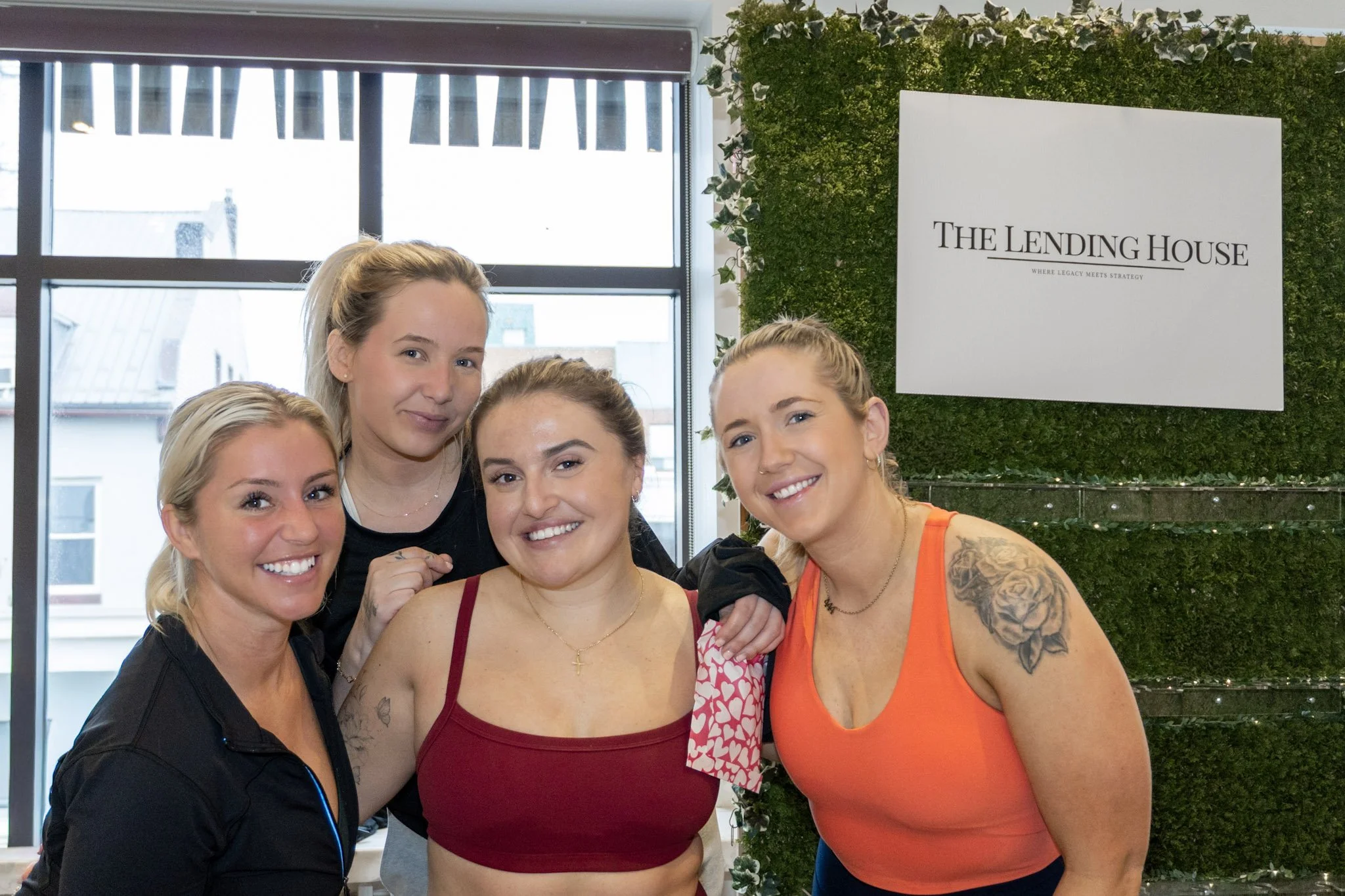 Four smiling women posing together indoors near a window with a sign reading 'The Lending House' on a green wall.