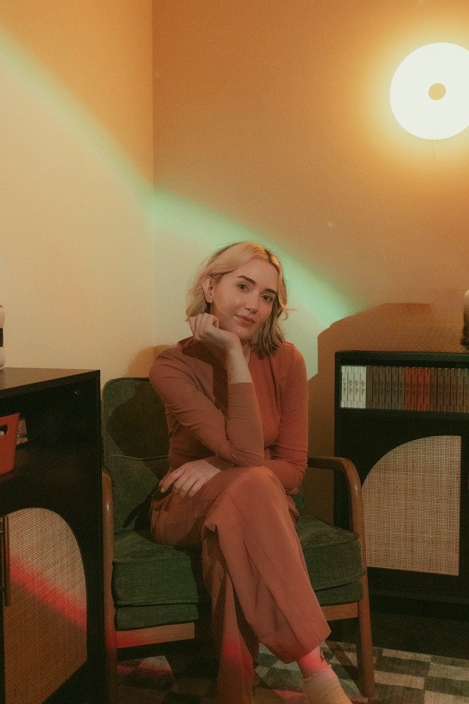 A woman with blonde hair sitting on a green cushioned chair in a cozy room with warm lighting, a bookshelf in the background, and colorful lighting effects on the wall.