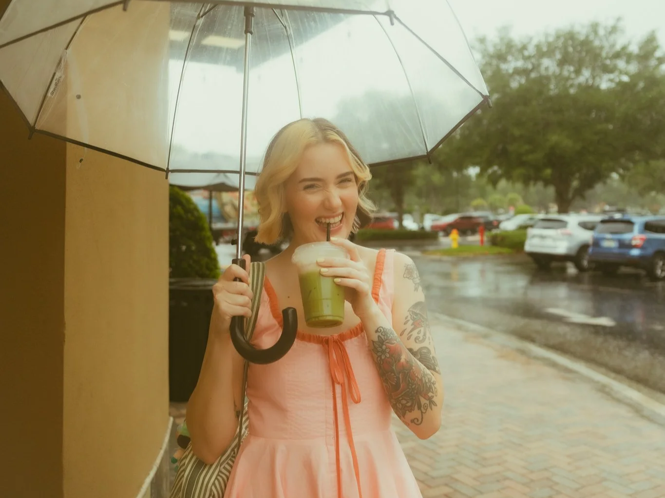 A young woman holding a clear umbrella and a green iced drink with a straw, standing outside on a rainy day, smiling and appearing happy.