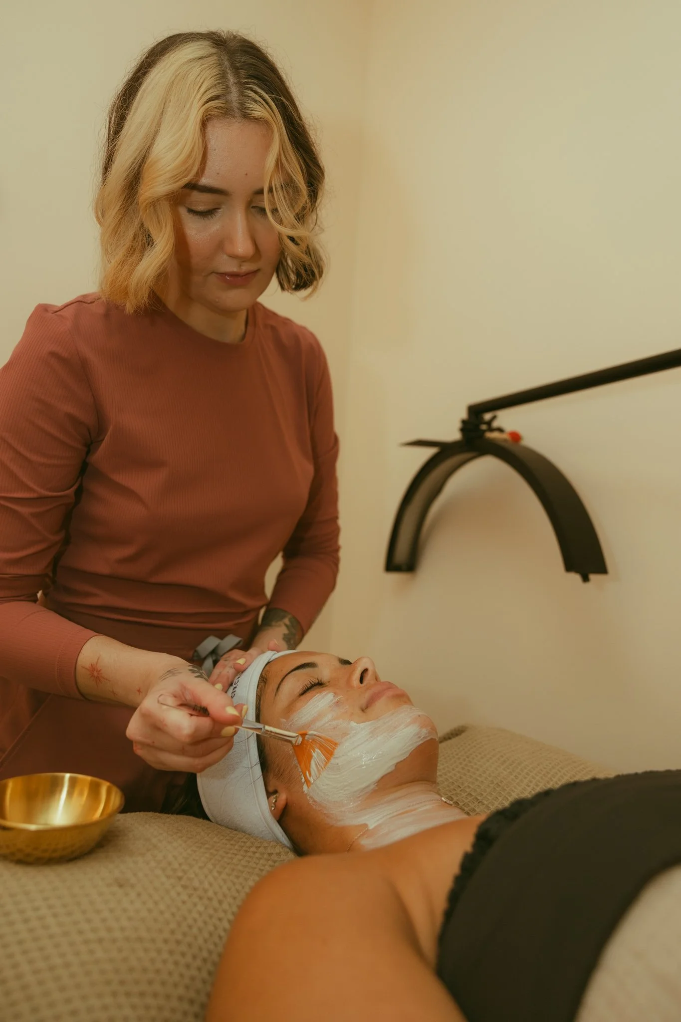 A woman receiving a facial treatment at a spa, lying down with a facial mask, while the esthetician applies a product to her face.