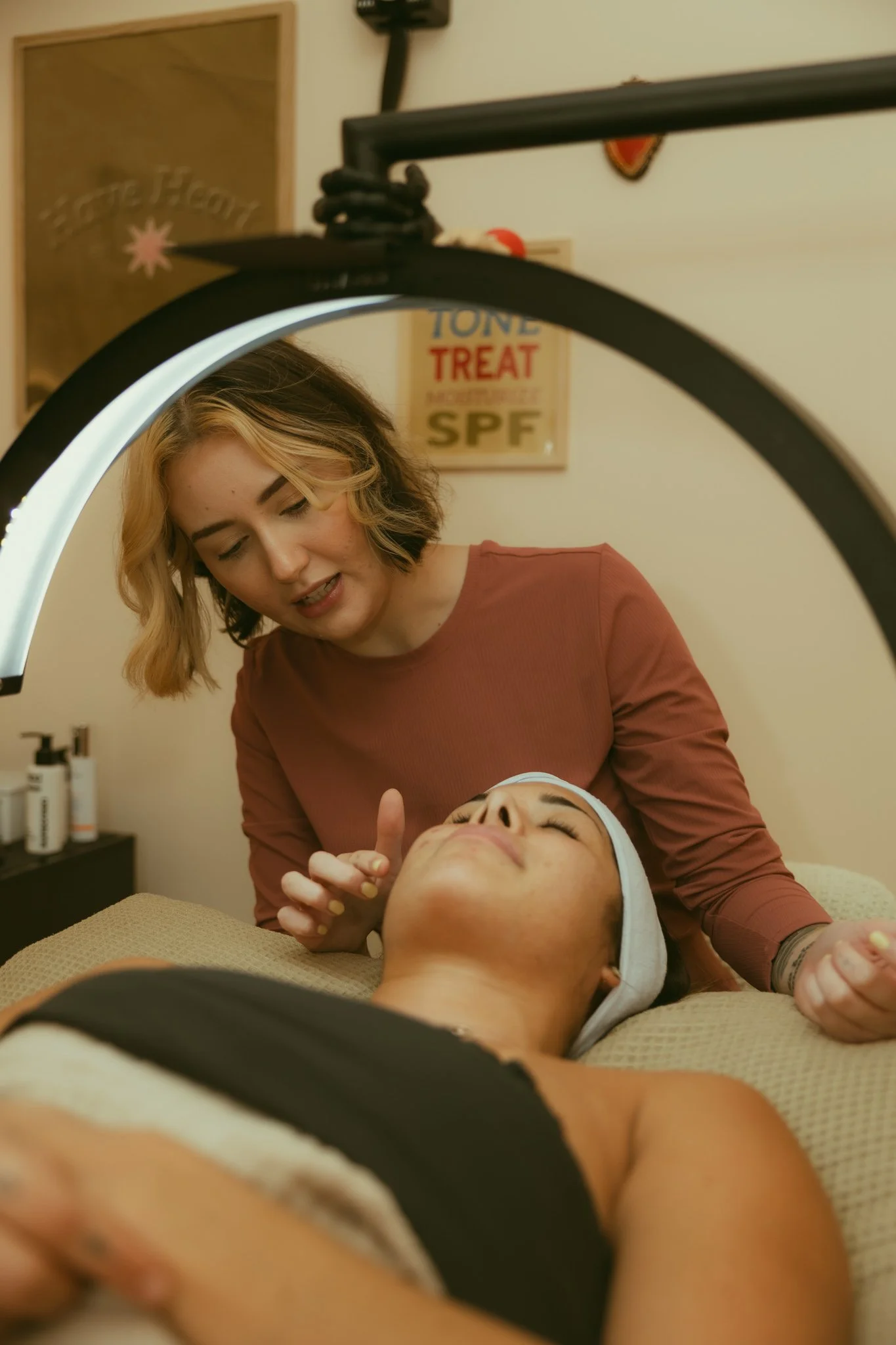 A woman providing facial treatment to a client lying on a treatment bed in a spa or skincare clinic. The woman is smiling and attentive, using her hand near the client's face. The client appears relaxed, with a towel wrapped around her head. The background features skincare products and decor.