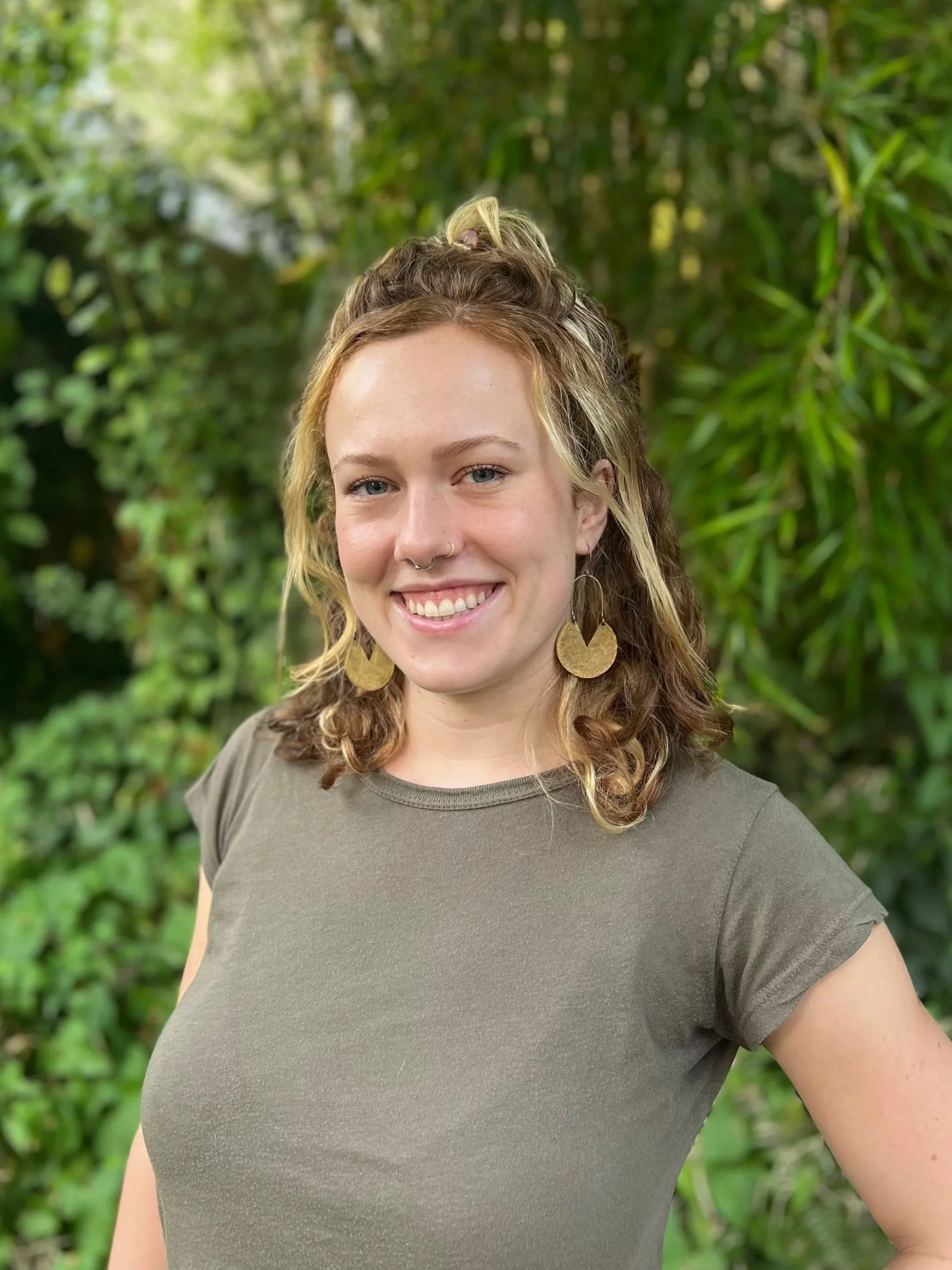 A young woman smiling outdoors with shoulder-length curly hair, wearing large gold disc earrings, a nose ring, and a gray t-shirt, standing in front of lush green foliage.