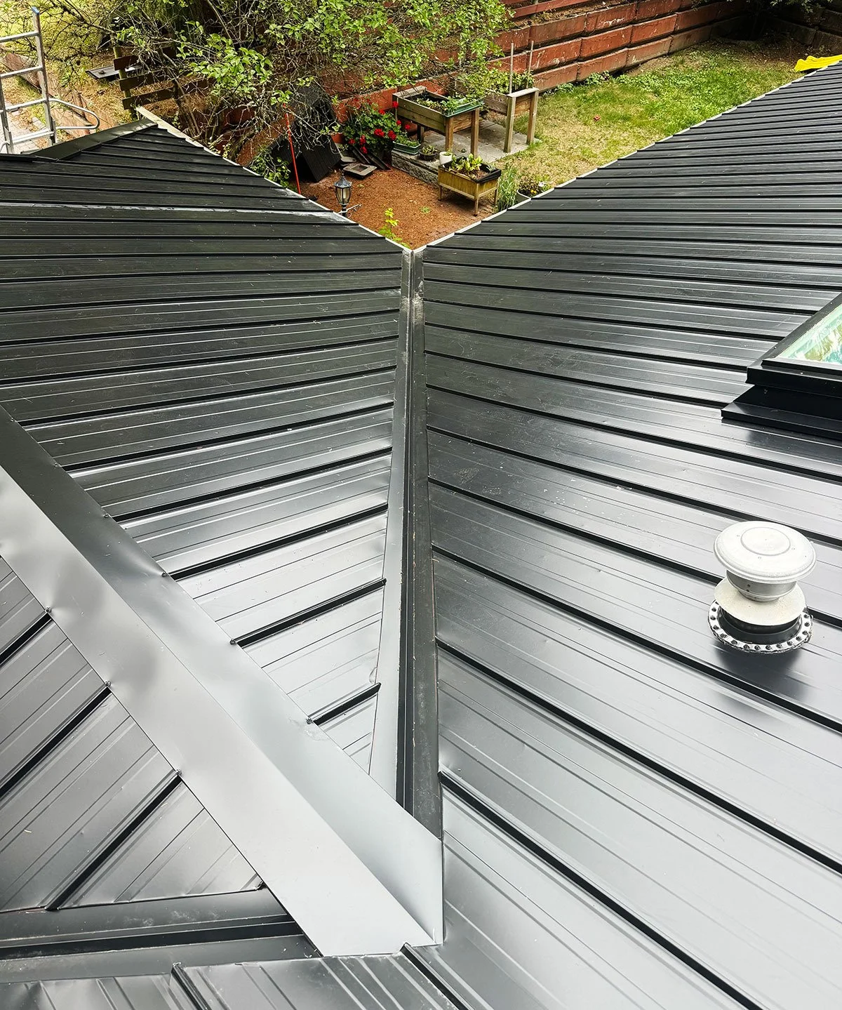 View of a metal roof with a steep pitch, showing the seam at the ridge, a vent pipe, and a small skylight, with a backyard featuring a brick wall, plants, and gardening boxes in the background.