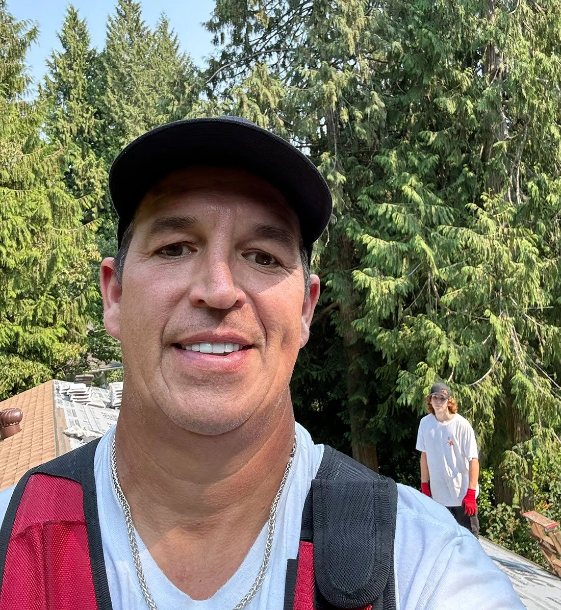 A man taking a selfie outdoors with a woman in the background, surrounded by tall green trees.