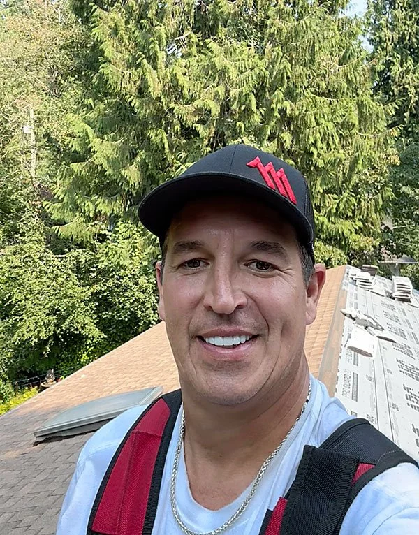 A man wearing a black cap with red lettering, a white shirt, and a backpack, smiling and standing outdoors on a roof with a background of green pine trees.